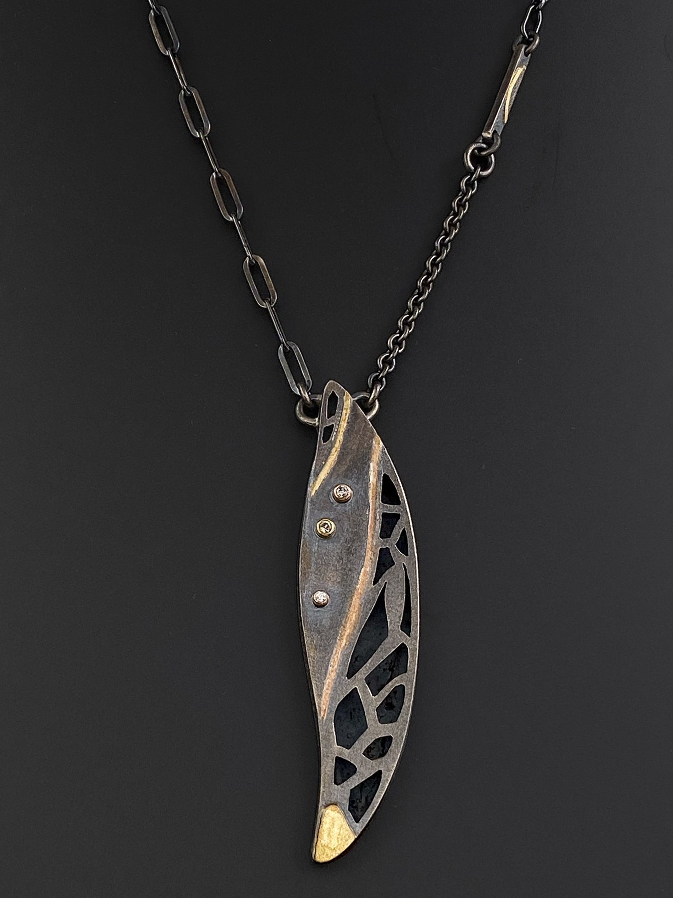Front view of an artisan mixed-metal feather pendant featuring intricate ajouré openwork, crafted in oxidized sterling silver with warm gold sweeps and diamond accents.