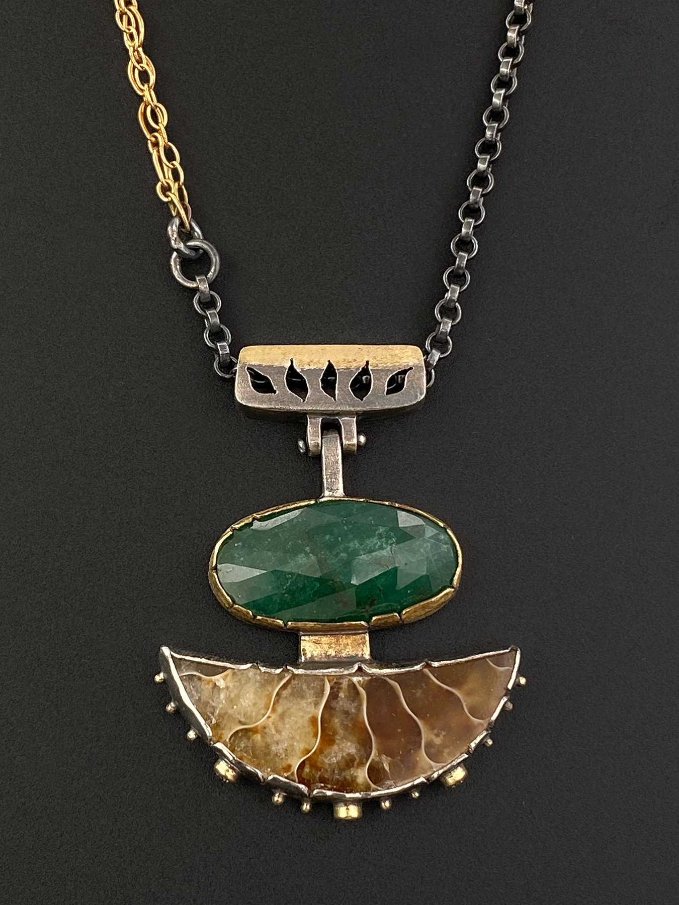 Front view of a one-of-a-kind talisman pendant featuring a faceted green emerald above a sliced brown ammonite fossil, set in 18k yellow gold and oxidized silver.