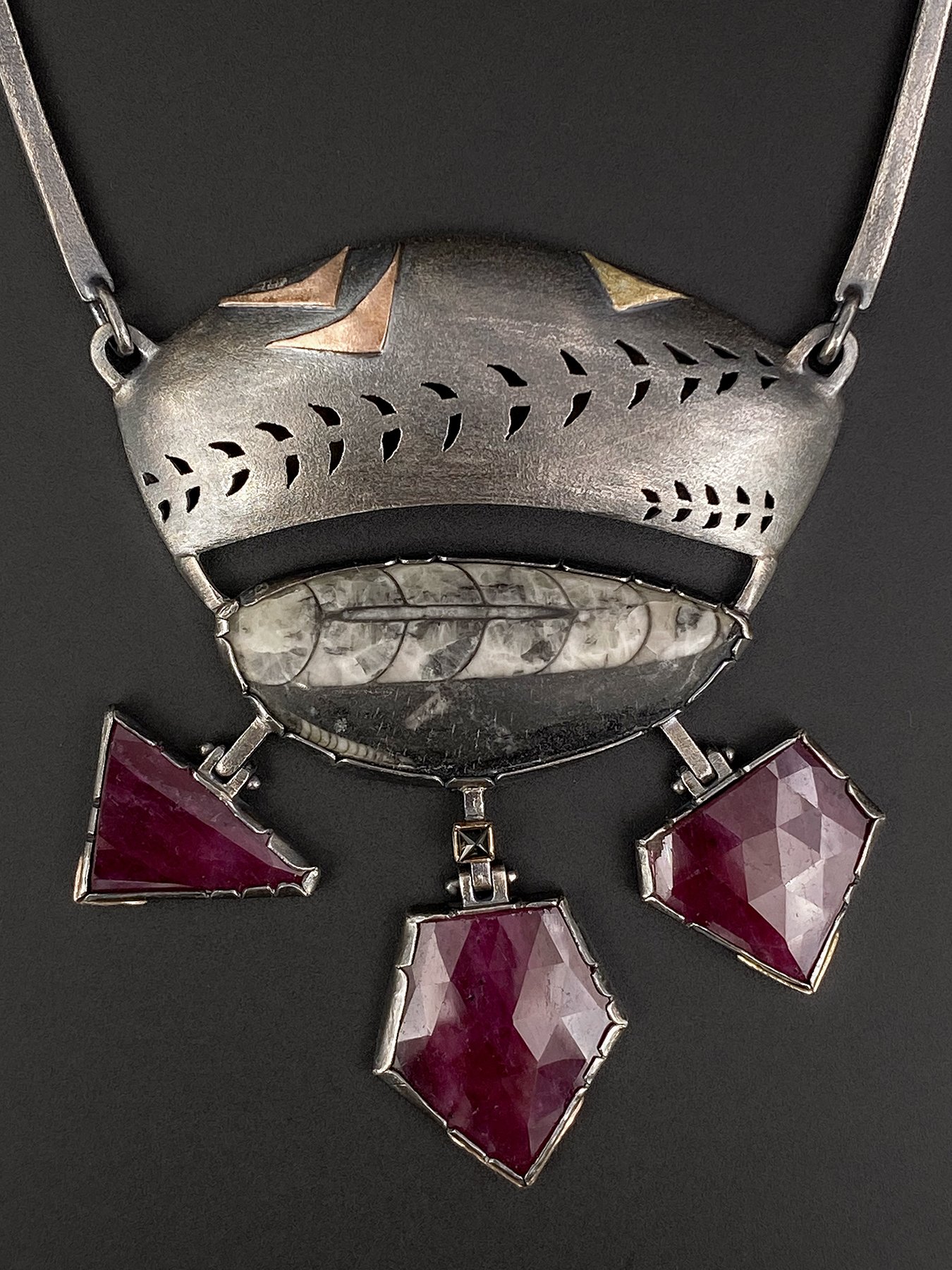 Front view of an architectural mixed metal statement necklace featuring a black and white Orthoceras fossil under a pierced oxidized silver canopy, with three hinged ruby drops.