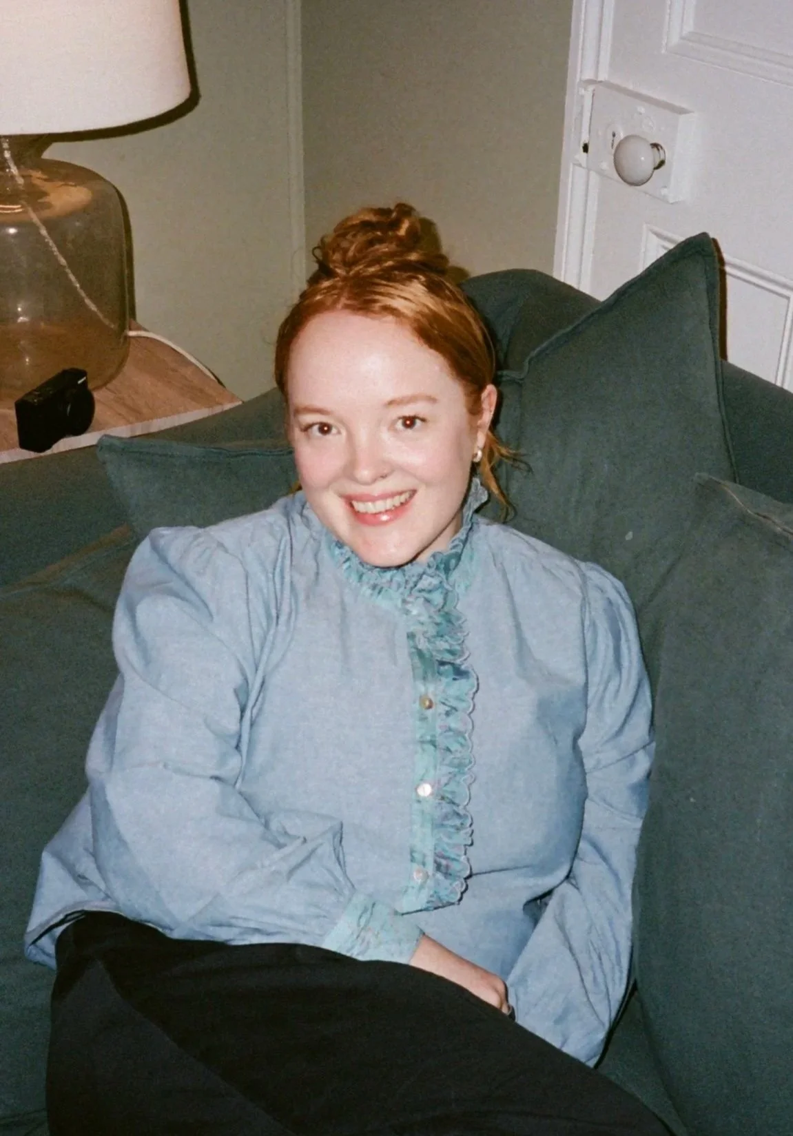 A smiling woman with red hair tied in a bun, sitting on a green couch with pillows, in a room with a lamp on a side table and a white door nearby.