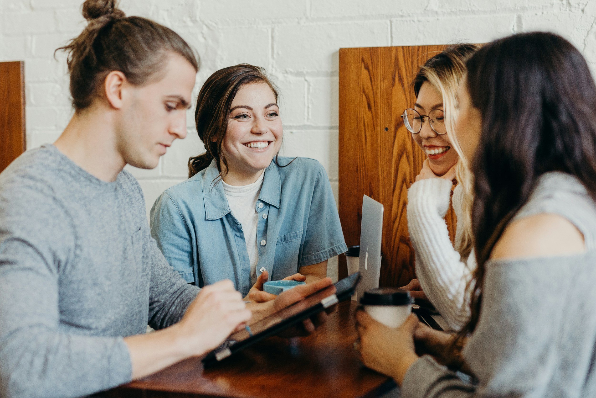 Three young women and a man sitting around a wooden table, engaging with one another and a tablet, in a cozy indoor setting with a white brick wall and wooden panels in the background.