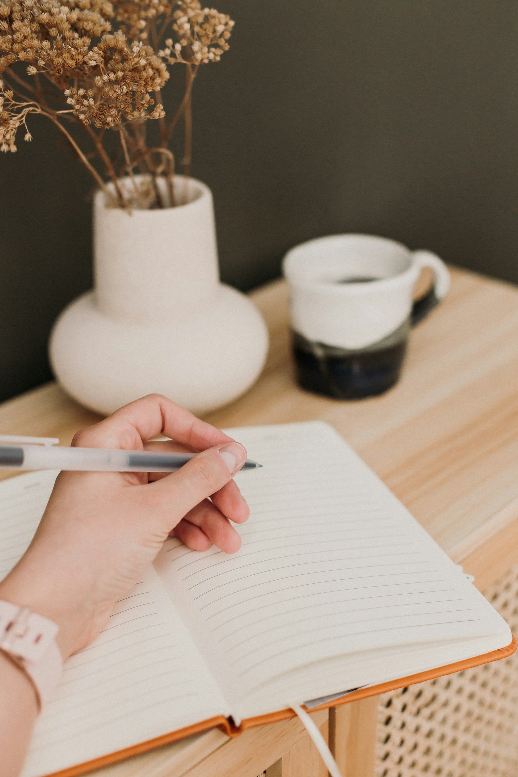 A person is writing in a notebook with a black pen. In the background, there is a white vase with dried flowers and a two-toned ceramic mug on a wooden surface, against a dark wall.