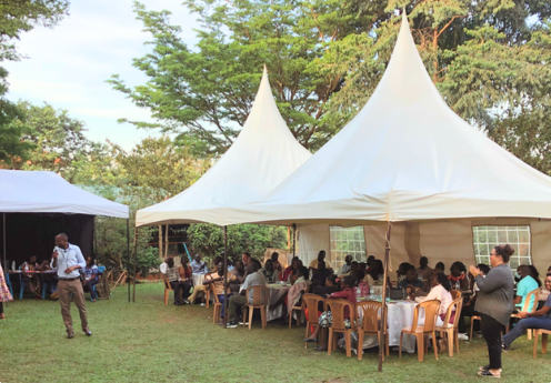 Outdoor event with people seated under white tents on a grassy area, surrounded by trees, during daytime.