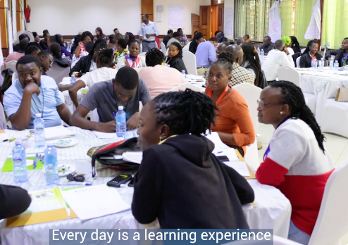 Group of people in a workshop setting at round tables, engaging in discussion and note-taking.