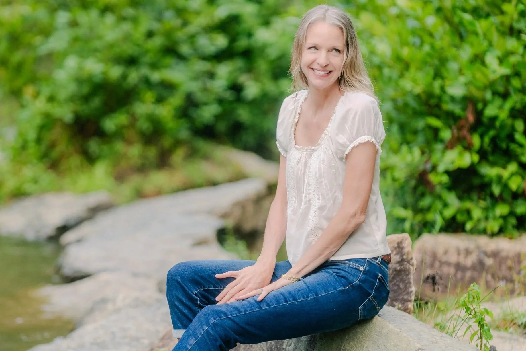 Dr. Lisa Robinson smiling, looking off to the side, and sitting on a rock next to a body of water surrounded by nature.