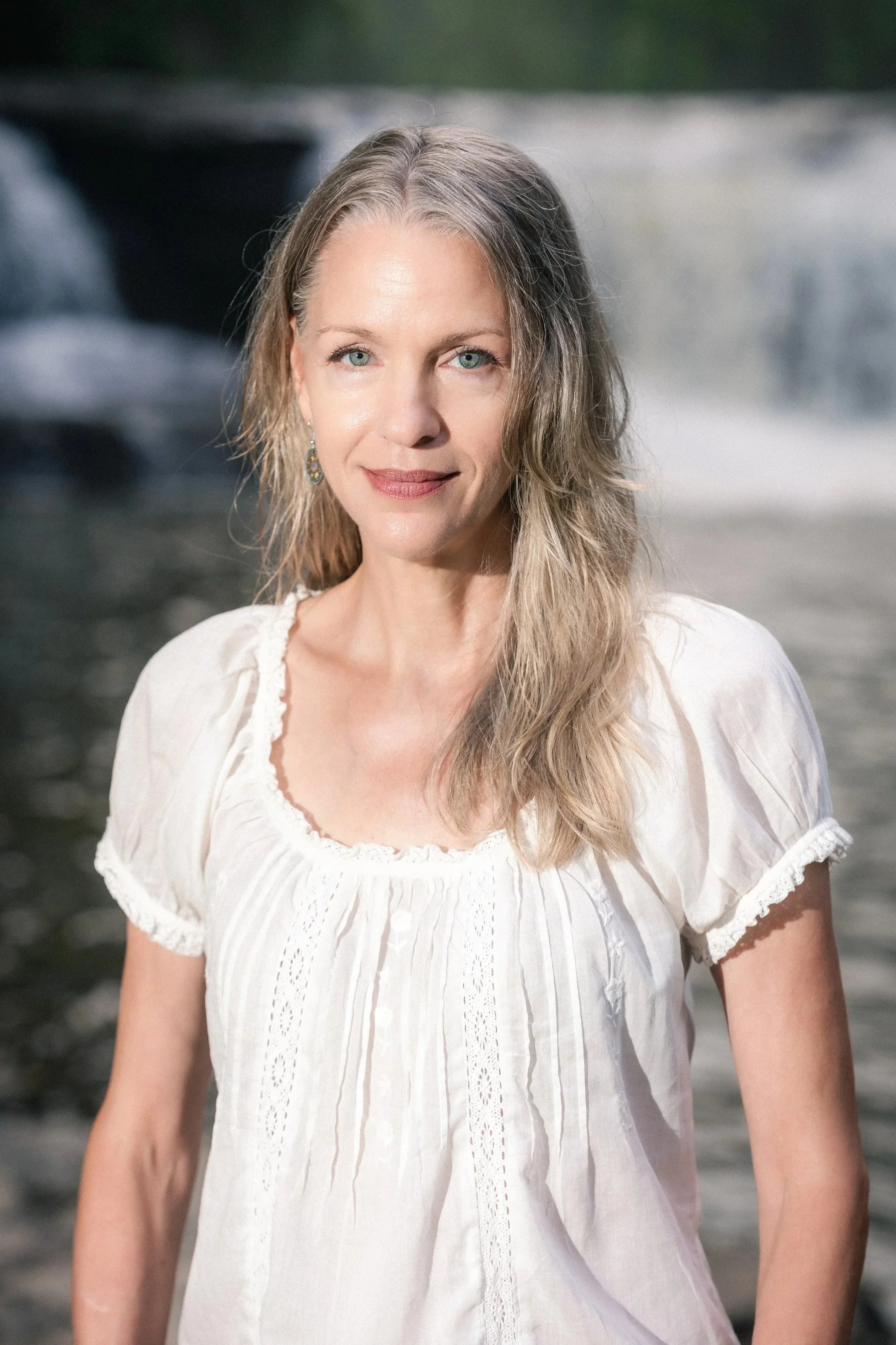 Dr. Lisa Robinson smiling standing outdoors in front of a body of water, wearing an off-white boho style top, with the water and waterfall blurred in the background.