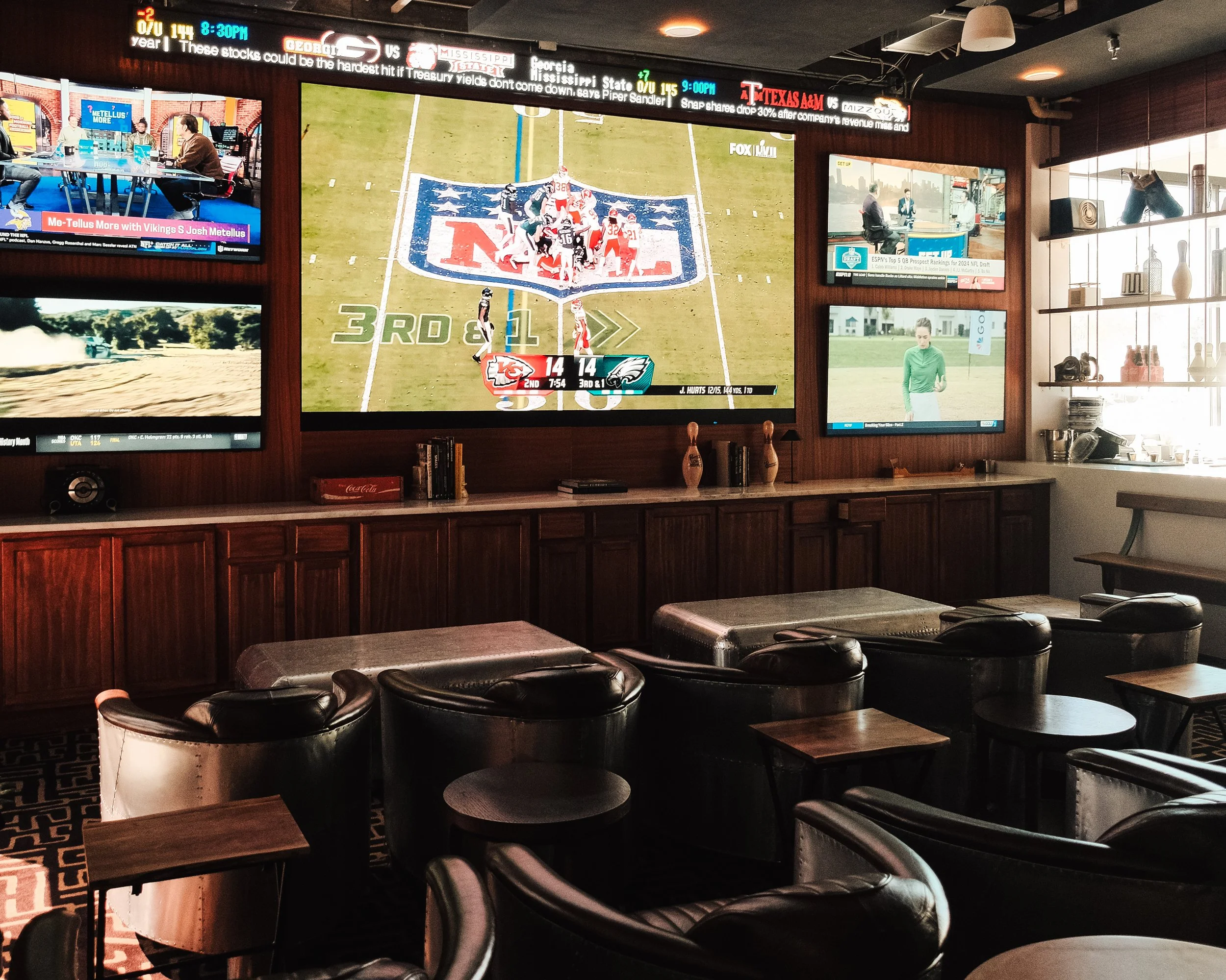 Sports bar interior with multiple TVs showing different channels, including a football game. Leather chairs and tables are arranged for viewing. Shelves with decorative items are on the right.