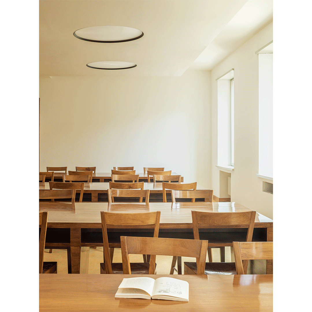 Aula vuota con tavoli e sedie di legno, finestra grande sulla parete laterale, due plafoniere rotonde sul soffitto, un libro aperto su uno dei tavoli.