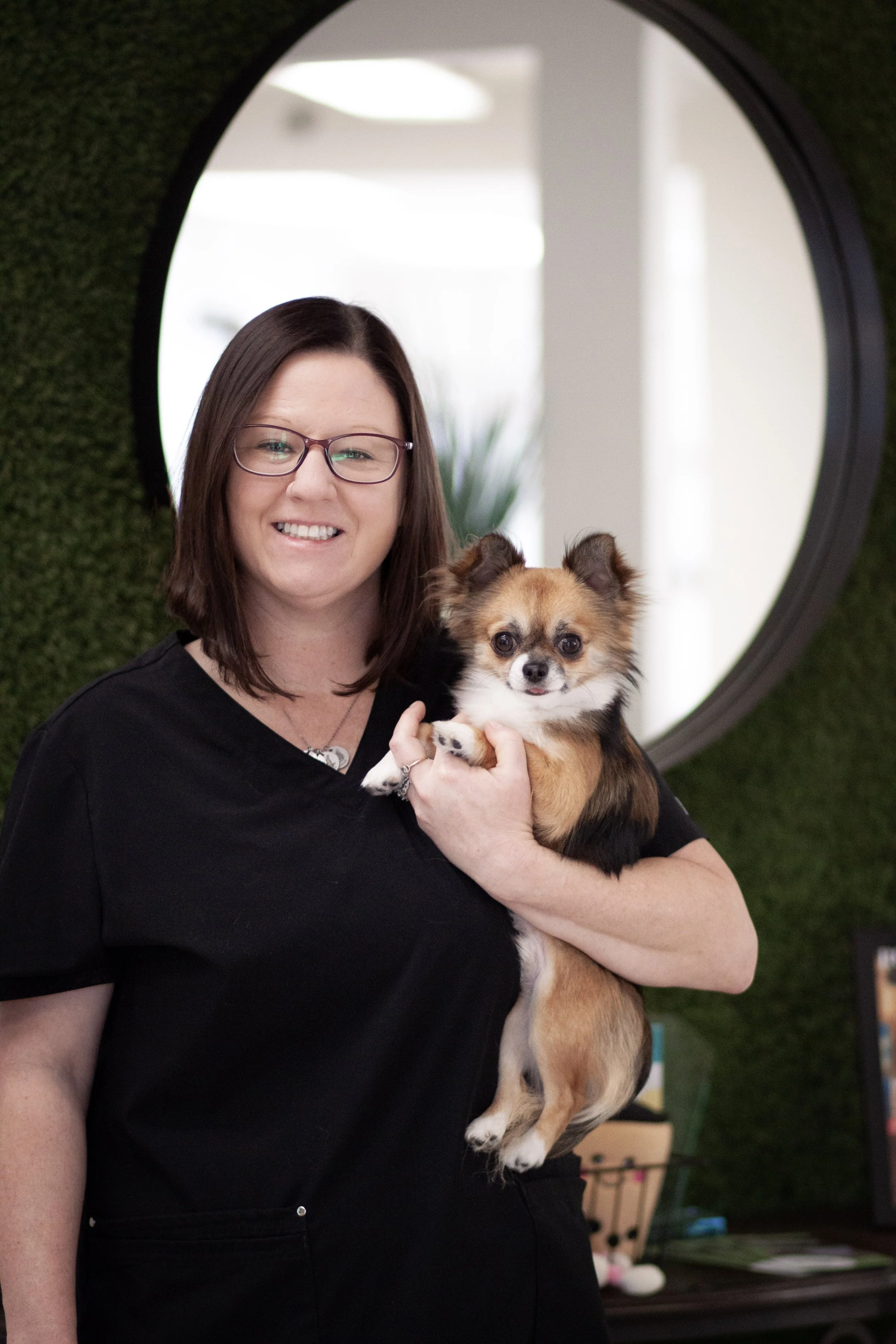 A woman with dark brown hair smiling and holding a small, fluffy white dog with light brown ears in front of a green wall with a round mirror.