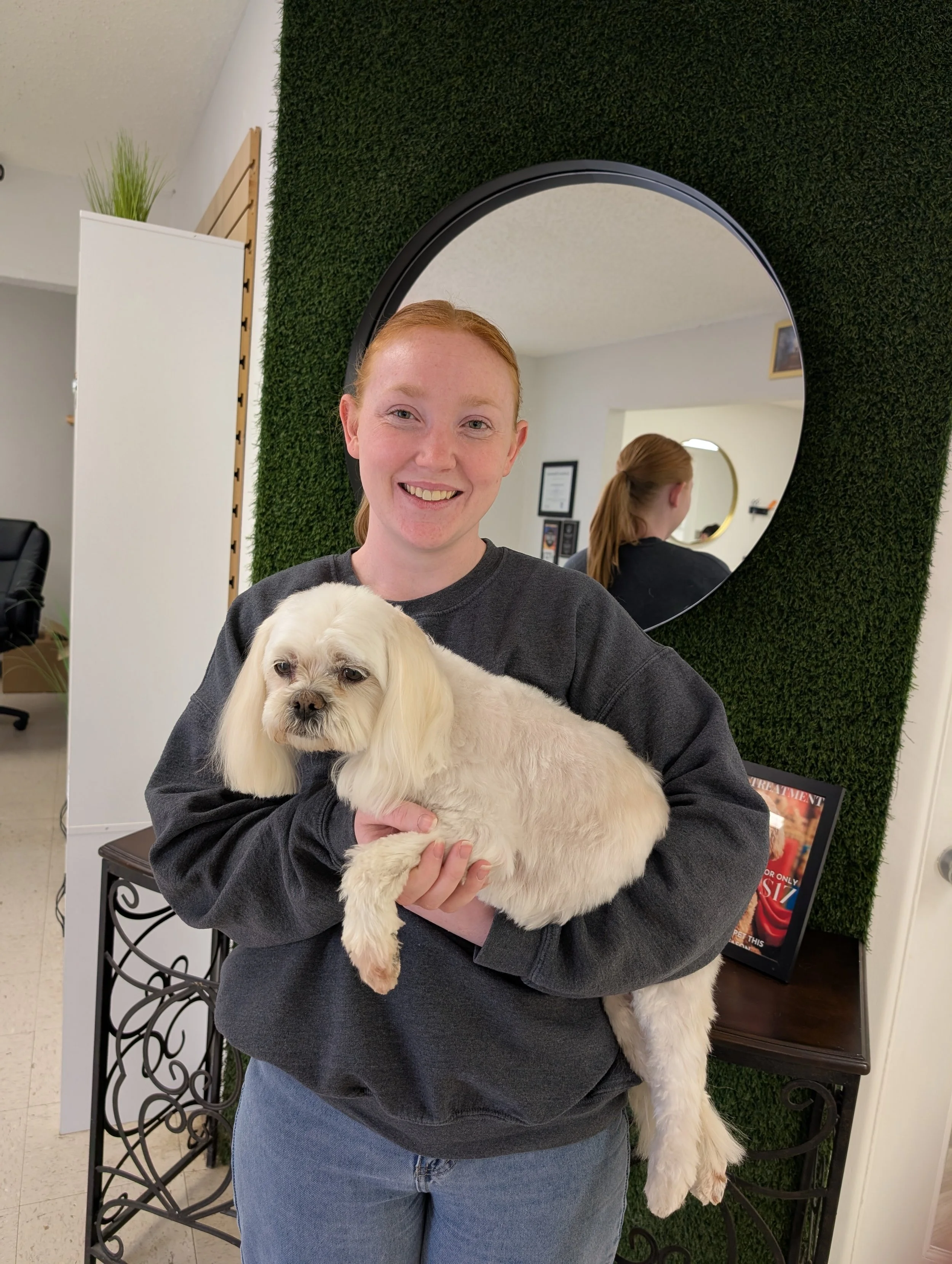 A woman with red hair, smiling and holding a small, white, fluffy dog in front of a green wall with a mirror reflecting her back and the dog.