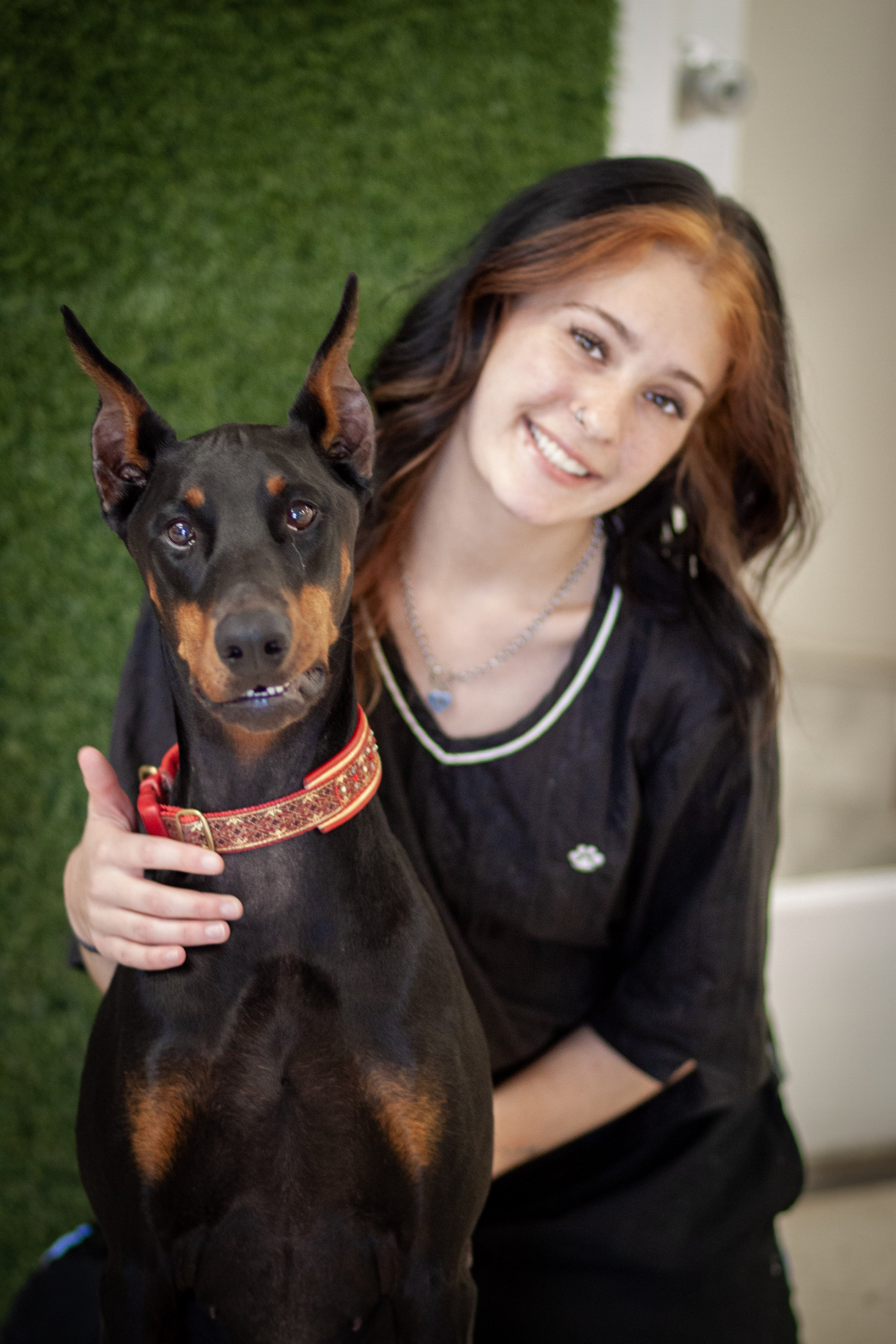 A young woman with long red hair holding a cream-colored dog with floppy ears and dark eyes, in front of a green wall with a round mirror.