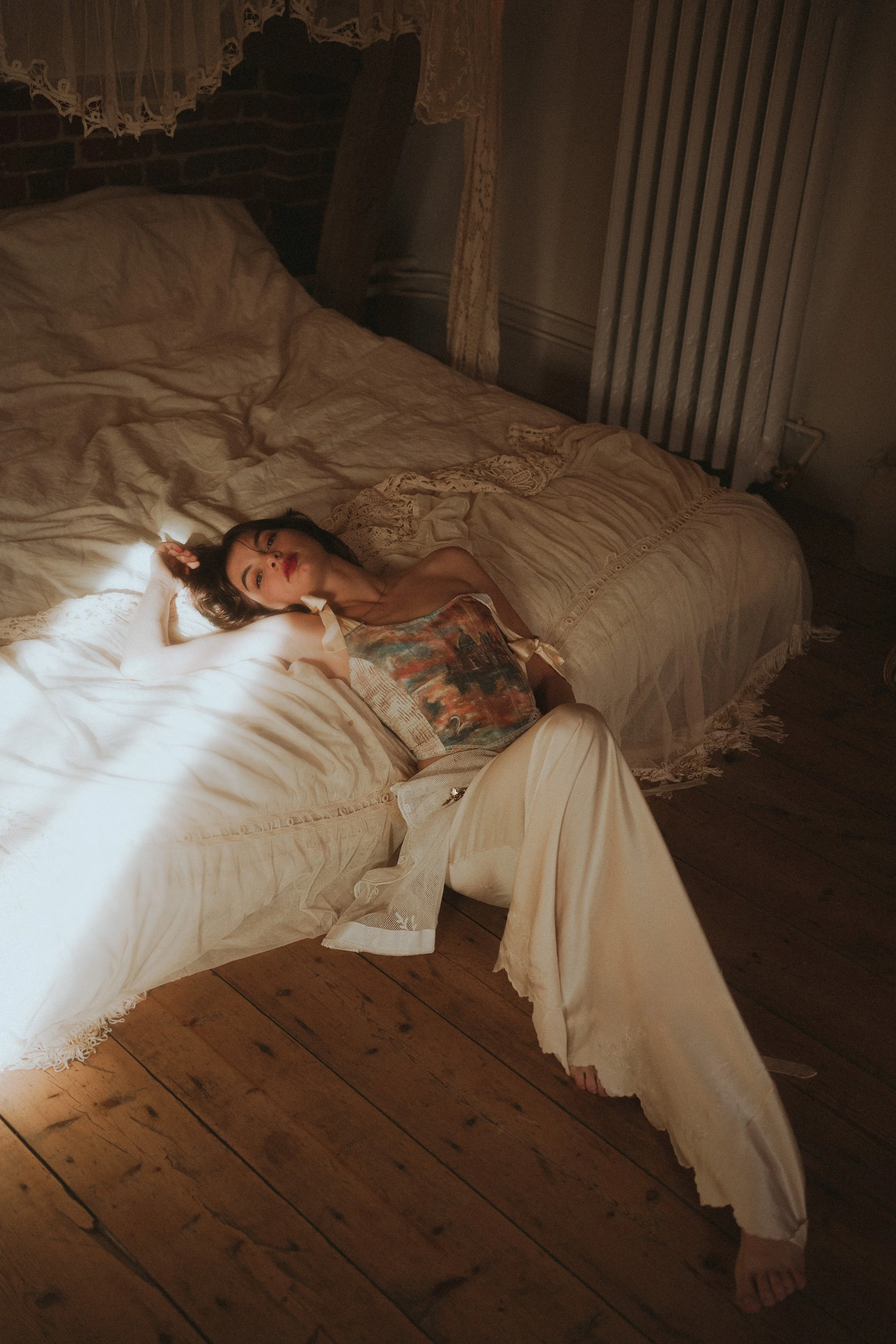 Une femme repose sur un sol en bois dur, à côté d'un lit à baldaquin, dans une pièce à l'ambiance tamisée et chaleureuse.