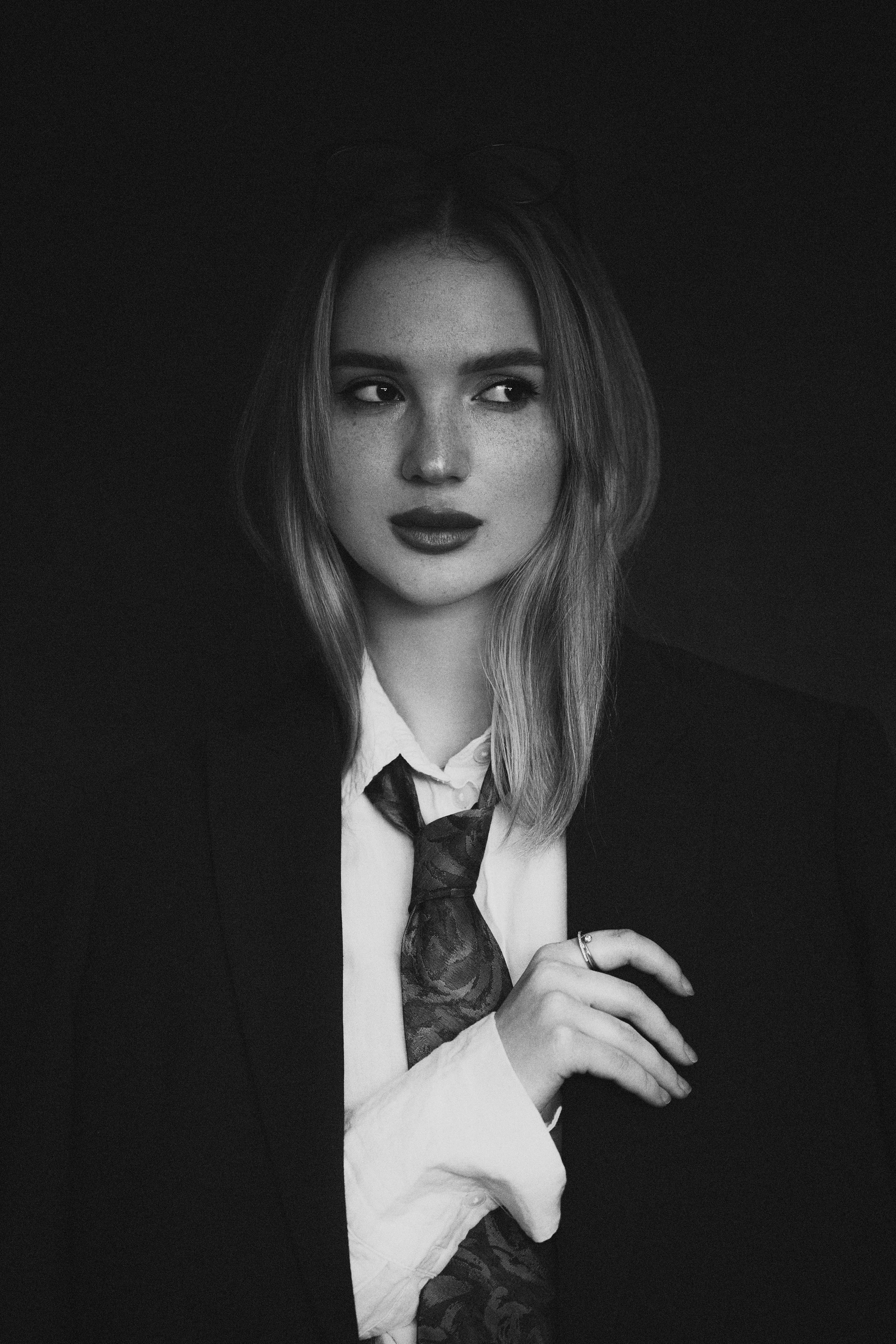 Portrait noir et blanc d'une femme avec des cheveux mi-longs, portant une chemise blanche, une veste noire et une cravate à motifs, tenant délicatement sa main avec une bague.