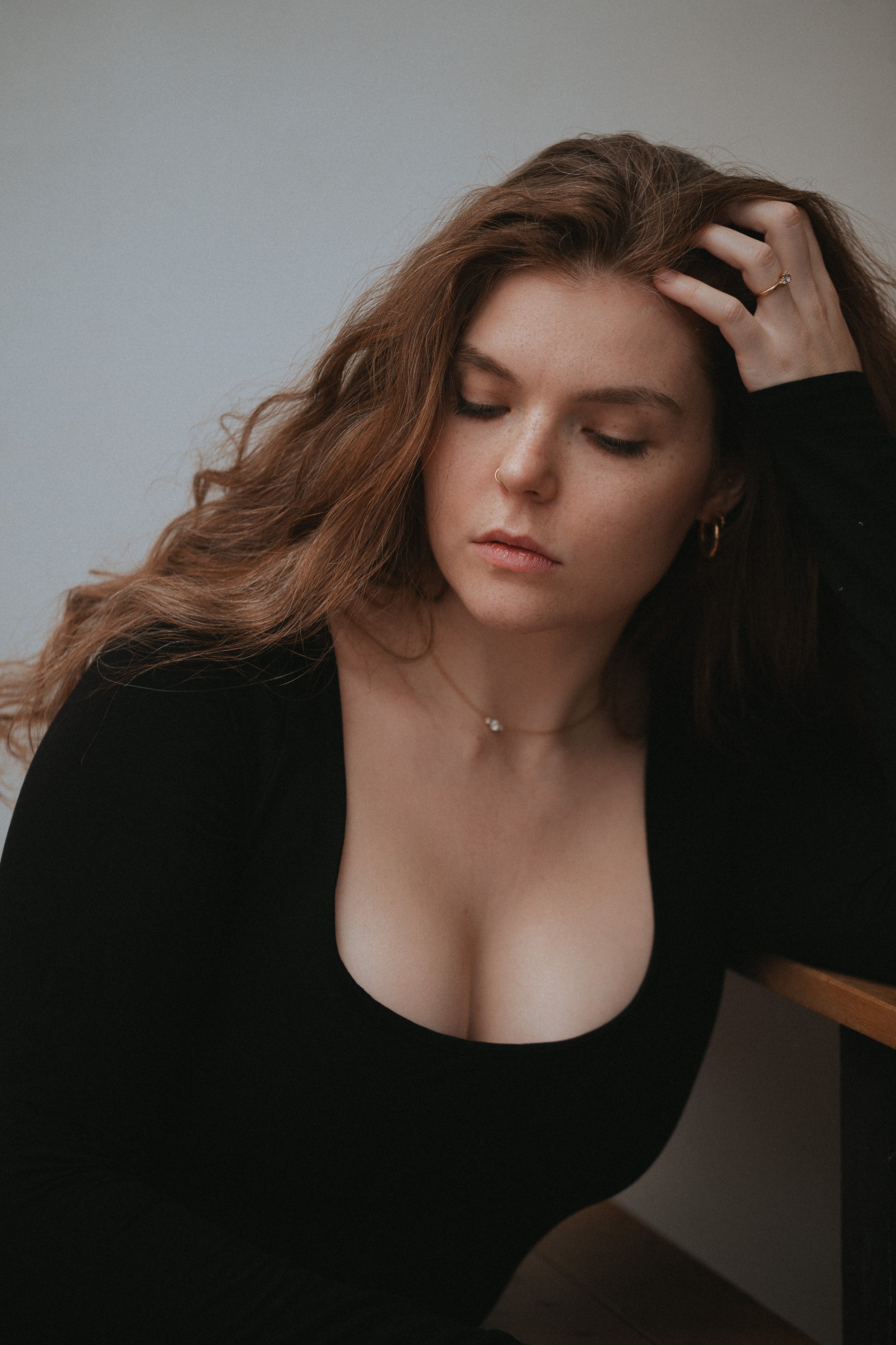 Jeune femme avec des cheveux bruns ondulés, portant un haut noir et des bijoux, les yeux fermés, appuyée contre un mur clair, avec une expression de contemplation.