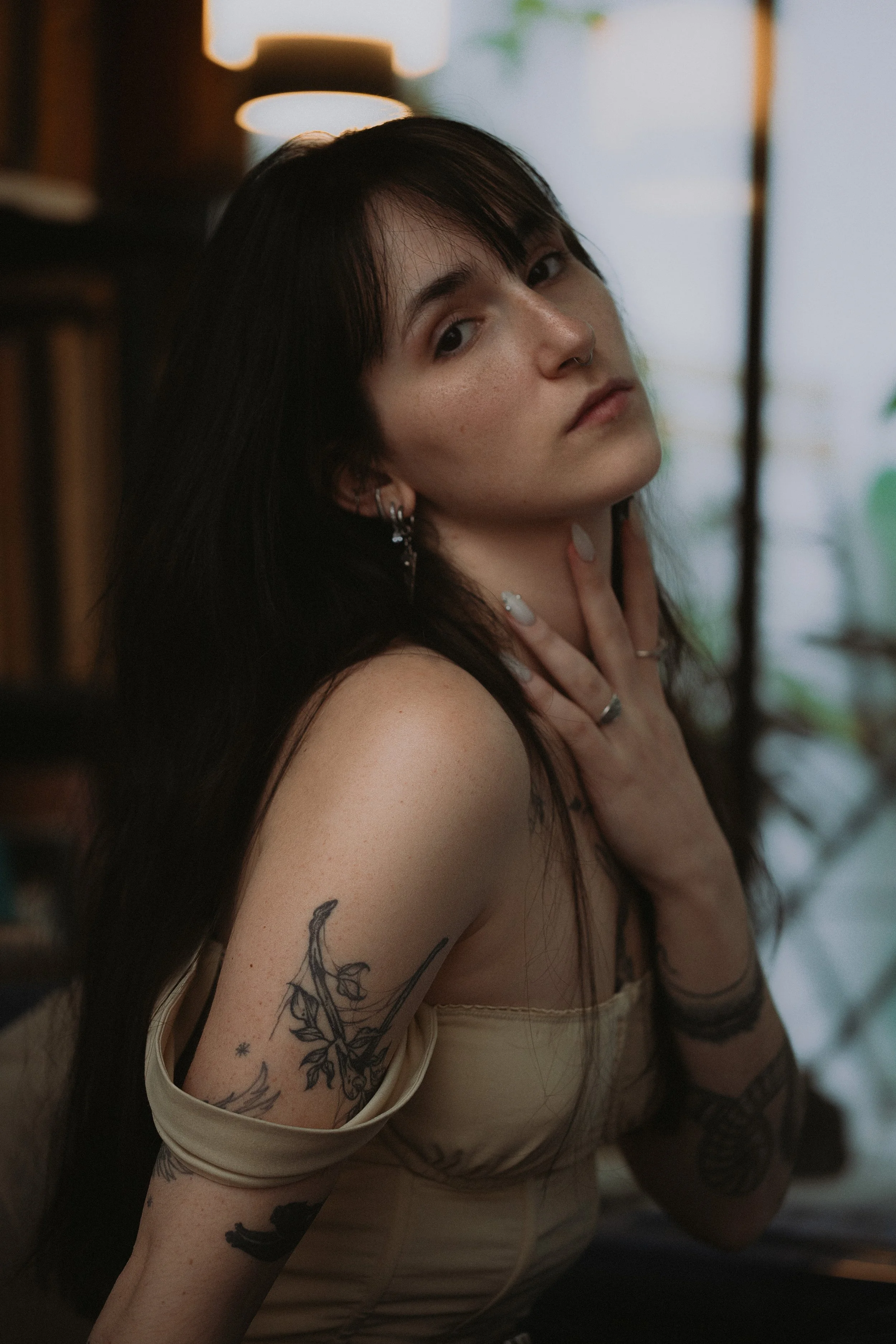 Une jeune femme aux cheveux longs et noirs, portant un haut beige, regarde fixement l'objectif, avec plusieurs tatouages visibles sur ses bras. Elle se tient dans un intérieur avec une lumière chaude et des plantes en arrière-plan.