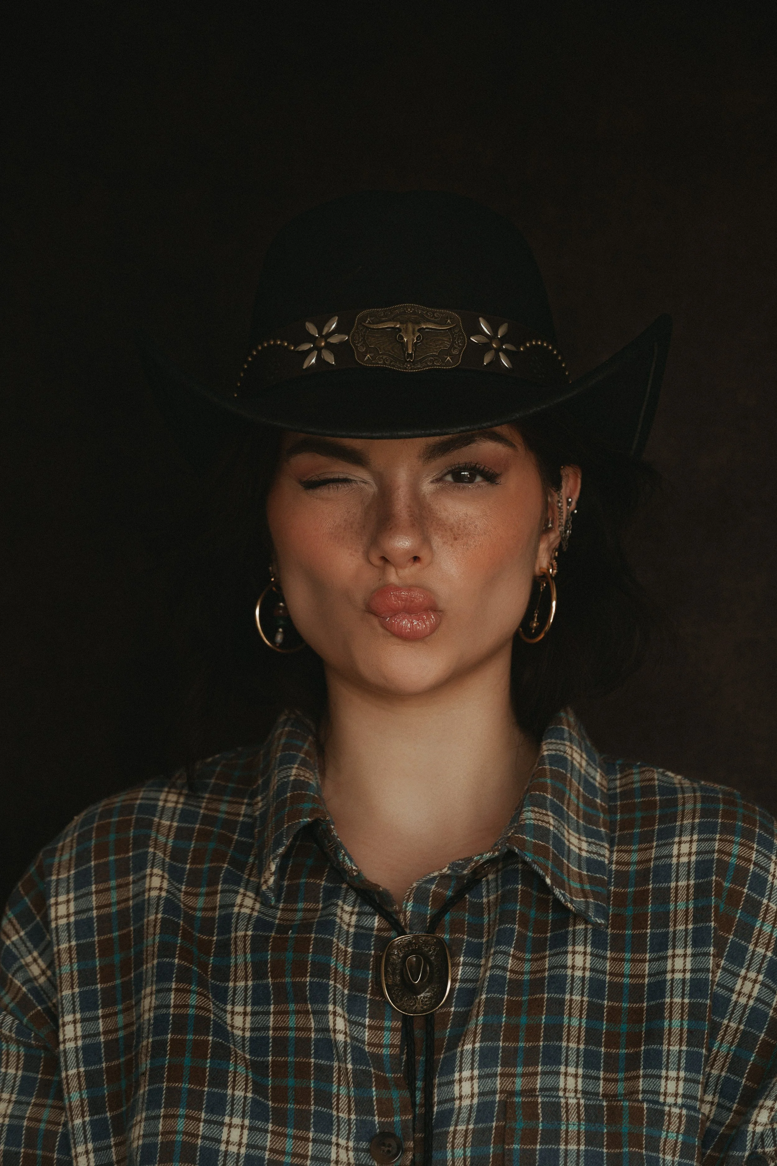 Une femme porte un chapeau de cowboy noir avec des décorations en métal, une chemise à carreaux, et des bijoux en or. Elle fait un clin d'œil et pince ses lèvres.