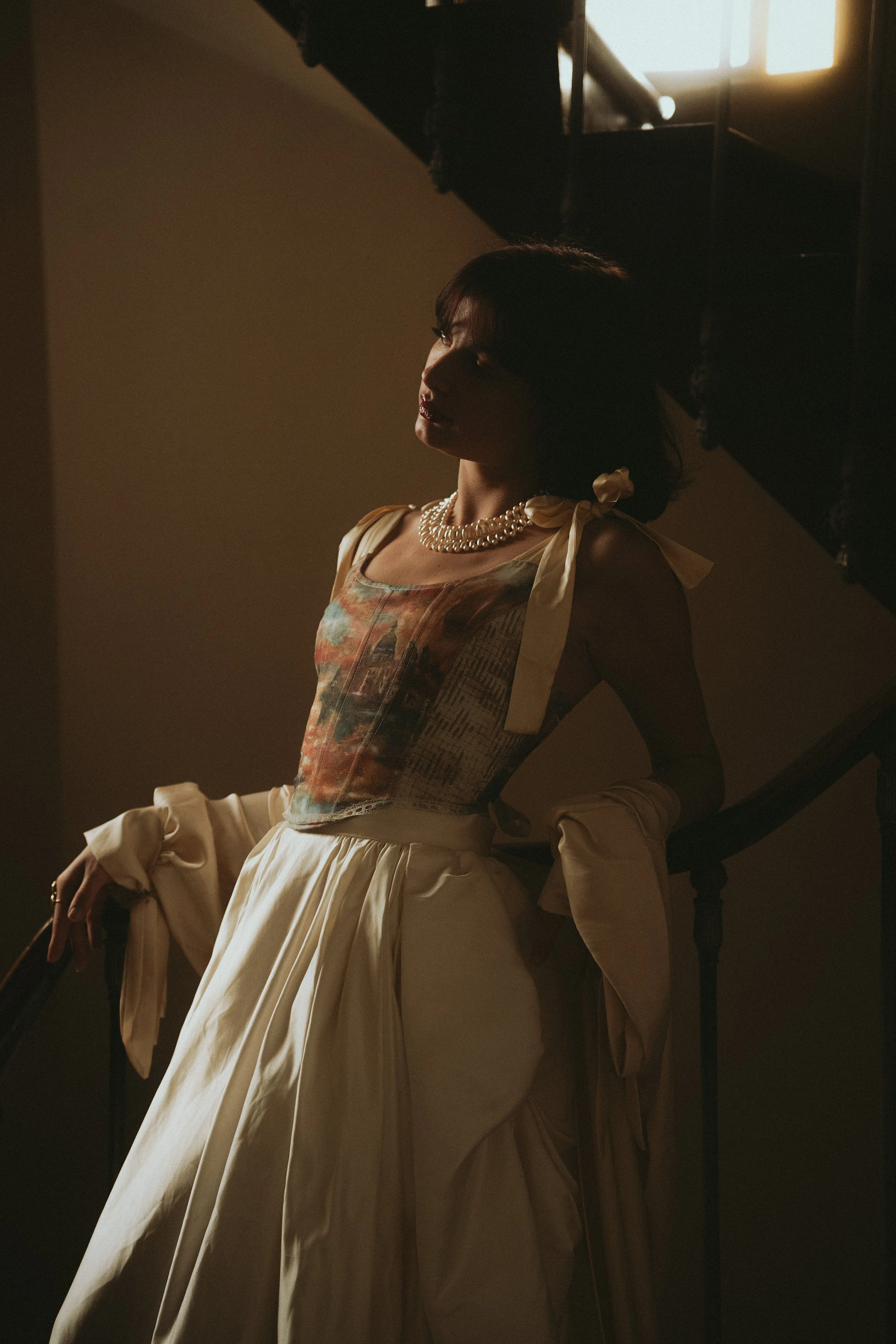 Femme élégante portant une robe longue avec un corsage coloré et une jupe beige, ornée d'un collier de perles et de manches bouffantes avec des rubans, se tenant sur un escalier dans une lumière tamisée.