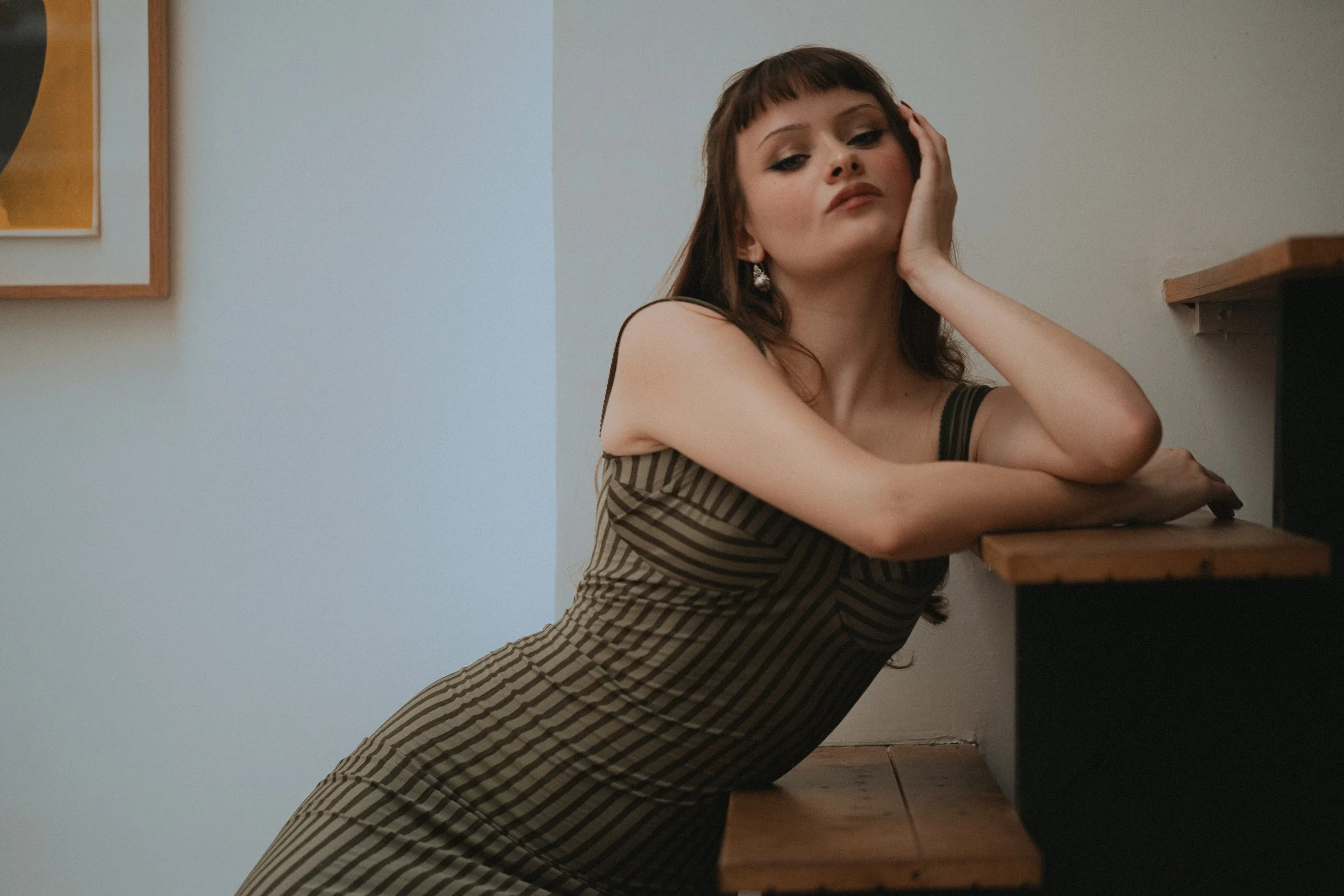 Jeune femme avec des cheveux longs et frisés, portant une robe à rayures noires et beiges, pose en appui sur une table en bois, regardant vers l'objectif.