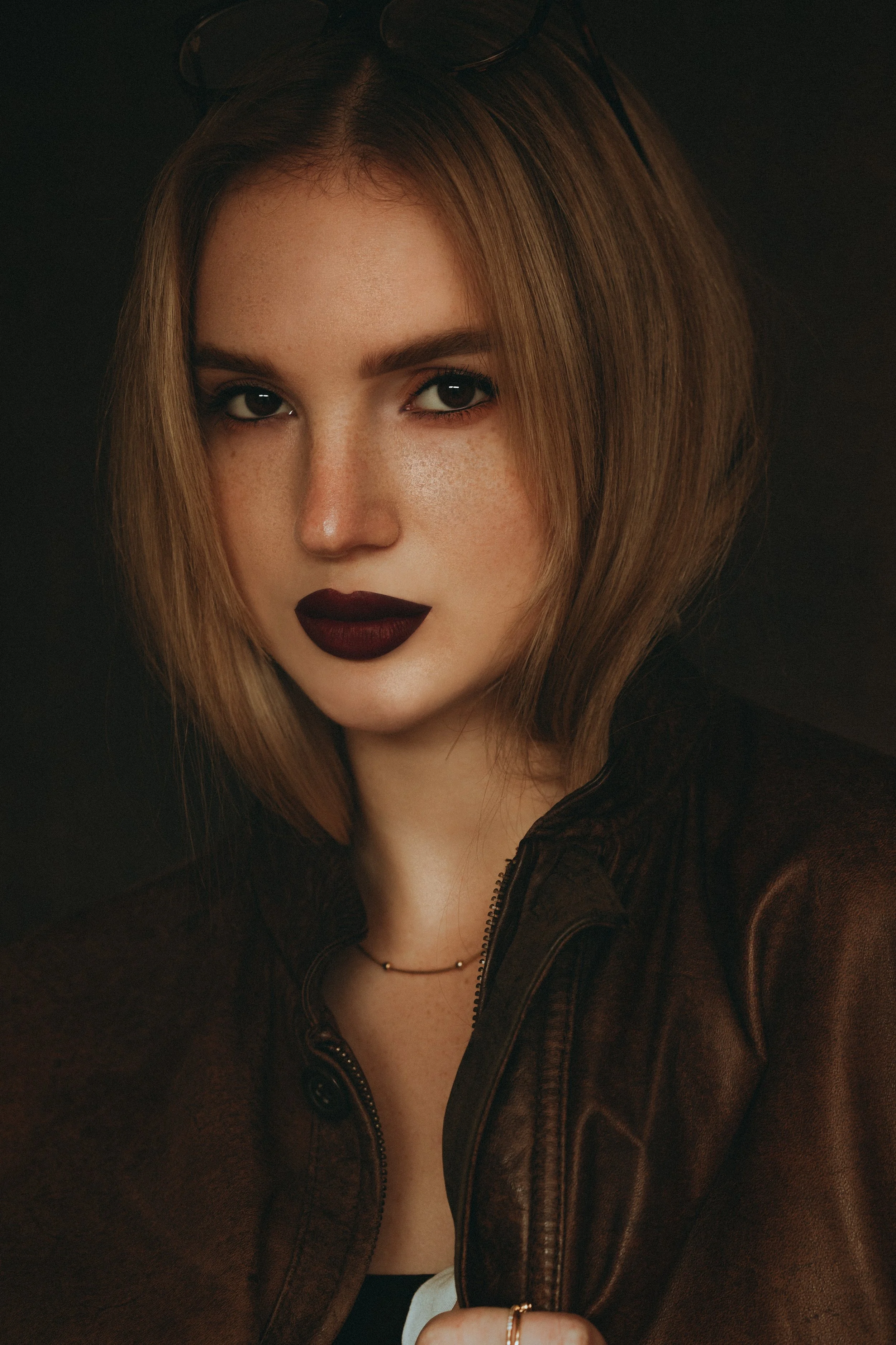 Jeune femme avec des cheveux bruns, maquillages foncés, portant une veste en cuir et une chaîne délicate, face à face, en studio avec fond sombre.