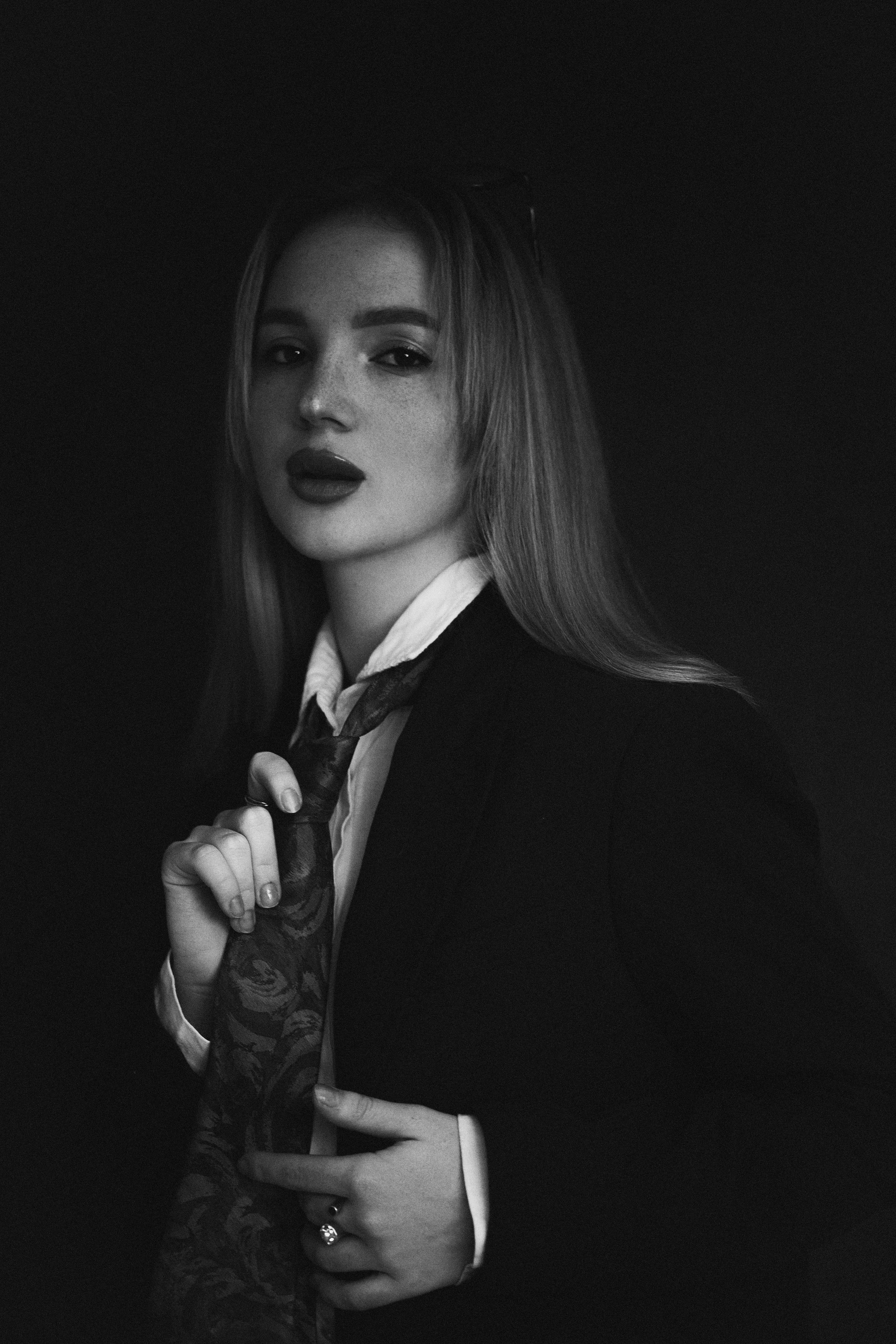 Une jeune femme aux cheveux longs, portant un costume noir et une cravate, pose sur fond noir, en noir et blanc.