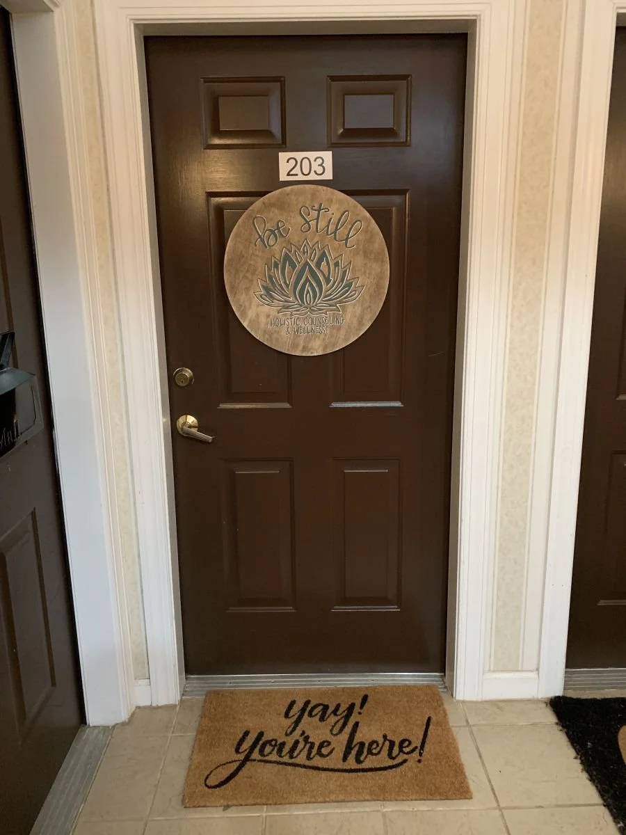 Interior hallway door for Suite 203 with a round Be Still Holistic Counseling and Wellness logo sign and a welcome mat reading yay you're here | Be Still Holistic Counseling & Wellness