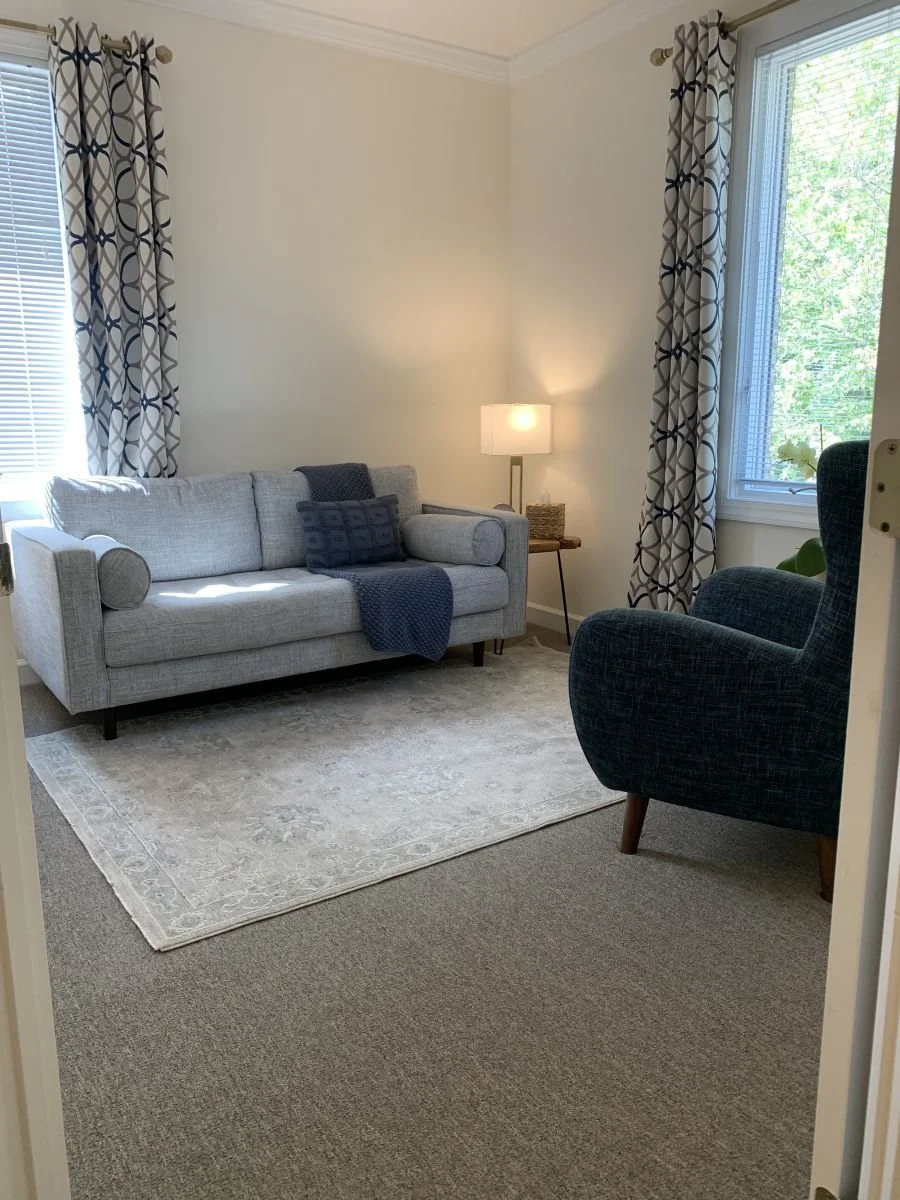 Cozy therapy office with a gray loveseat, navy accent chair, patterned curtains, and soft natural light | Be Still Holistic Counseling & Wellness