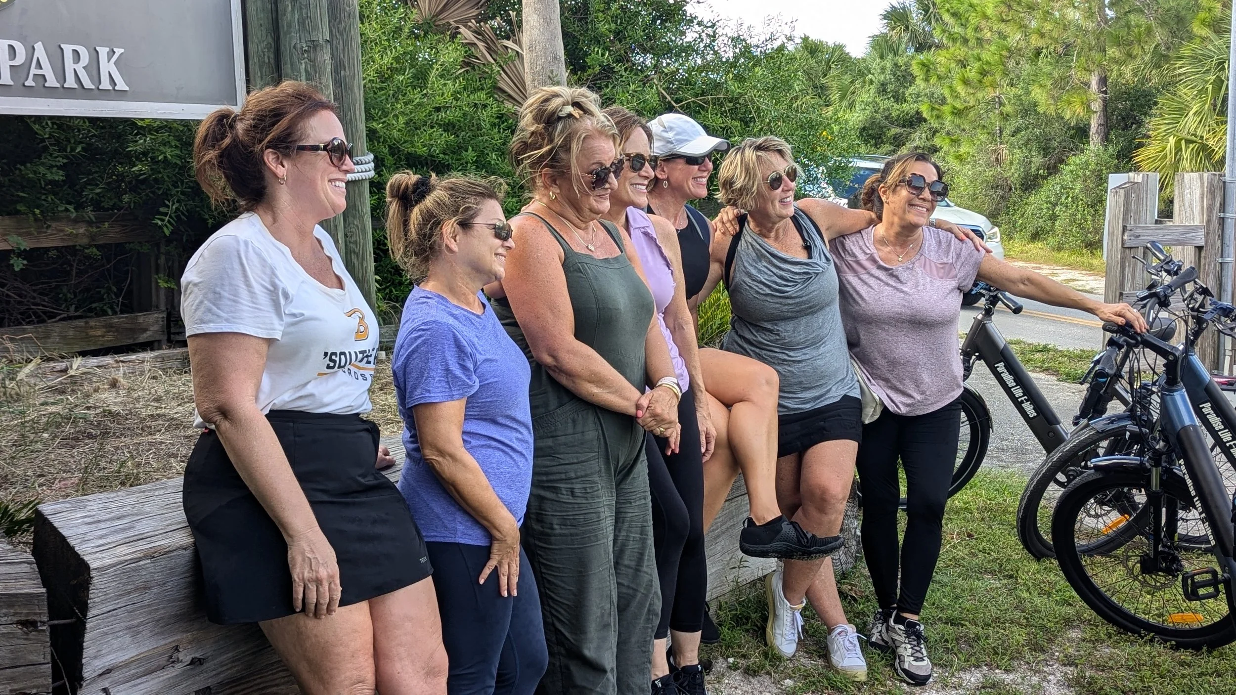 A group of seven women smiling and posing together outdoors near a wooden fence and trees. Some are wearing sunglasses, and there are electric bikes nearby.