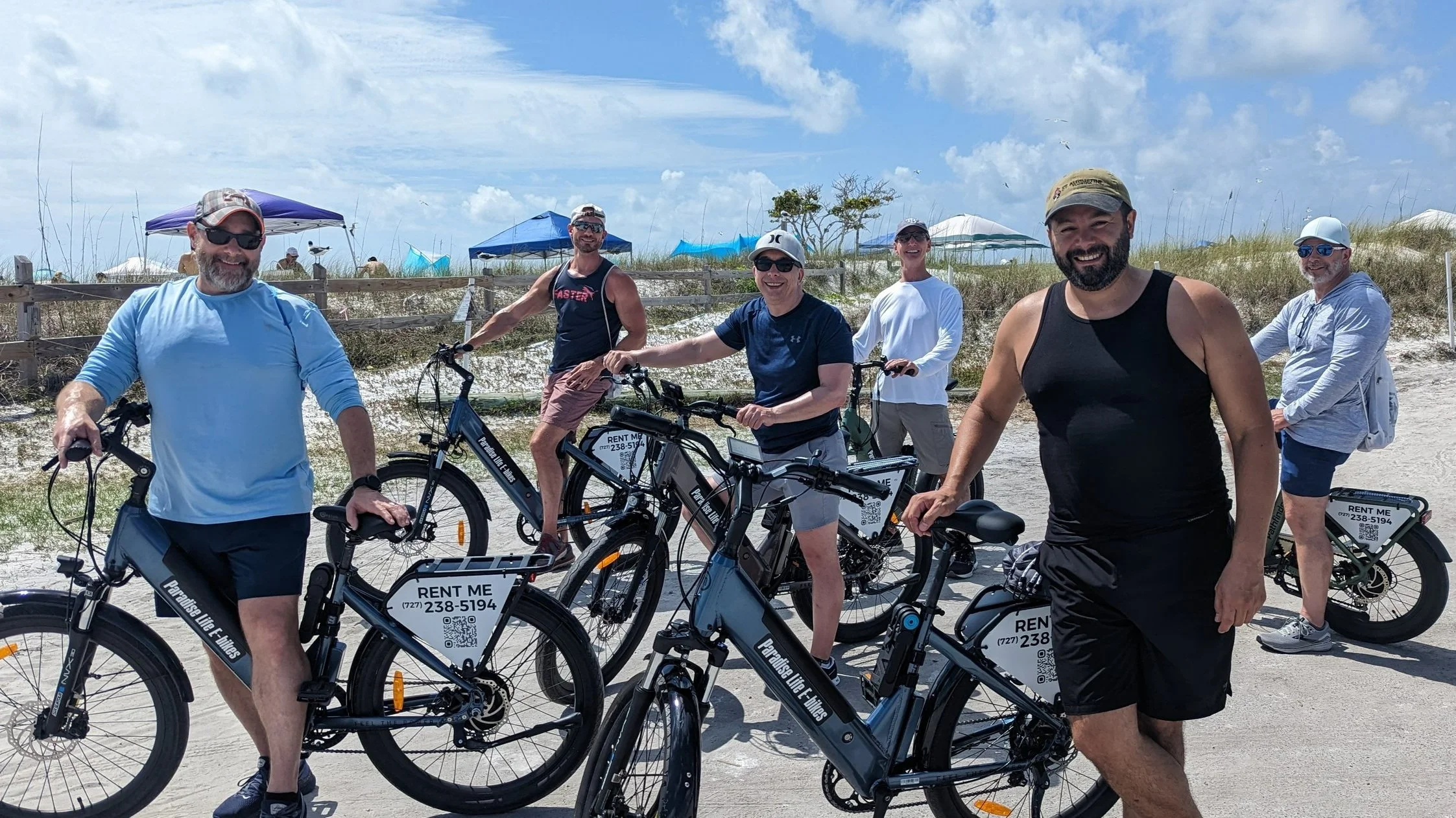Group of six men with bicycles on a sandy beach, smiling and wearing casual summer clothing, tents and a cloudy sky in the background.