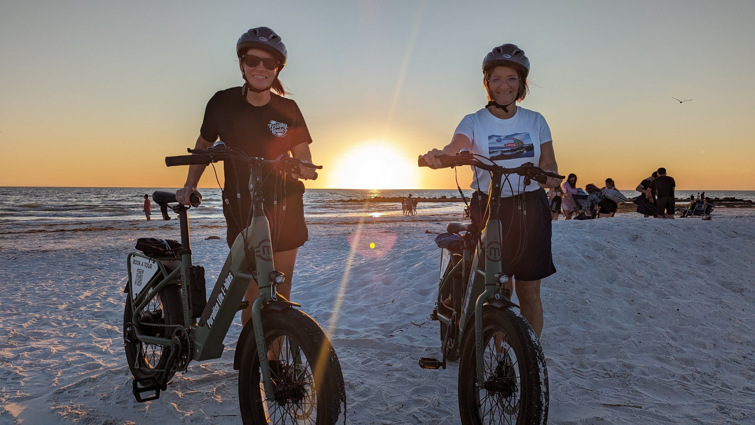 Two women with helmets and sunglasses standing next to electric bikes on a sandy beach at sunset, with other beachgoers in the background.