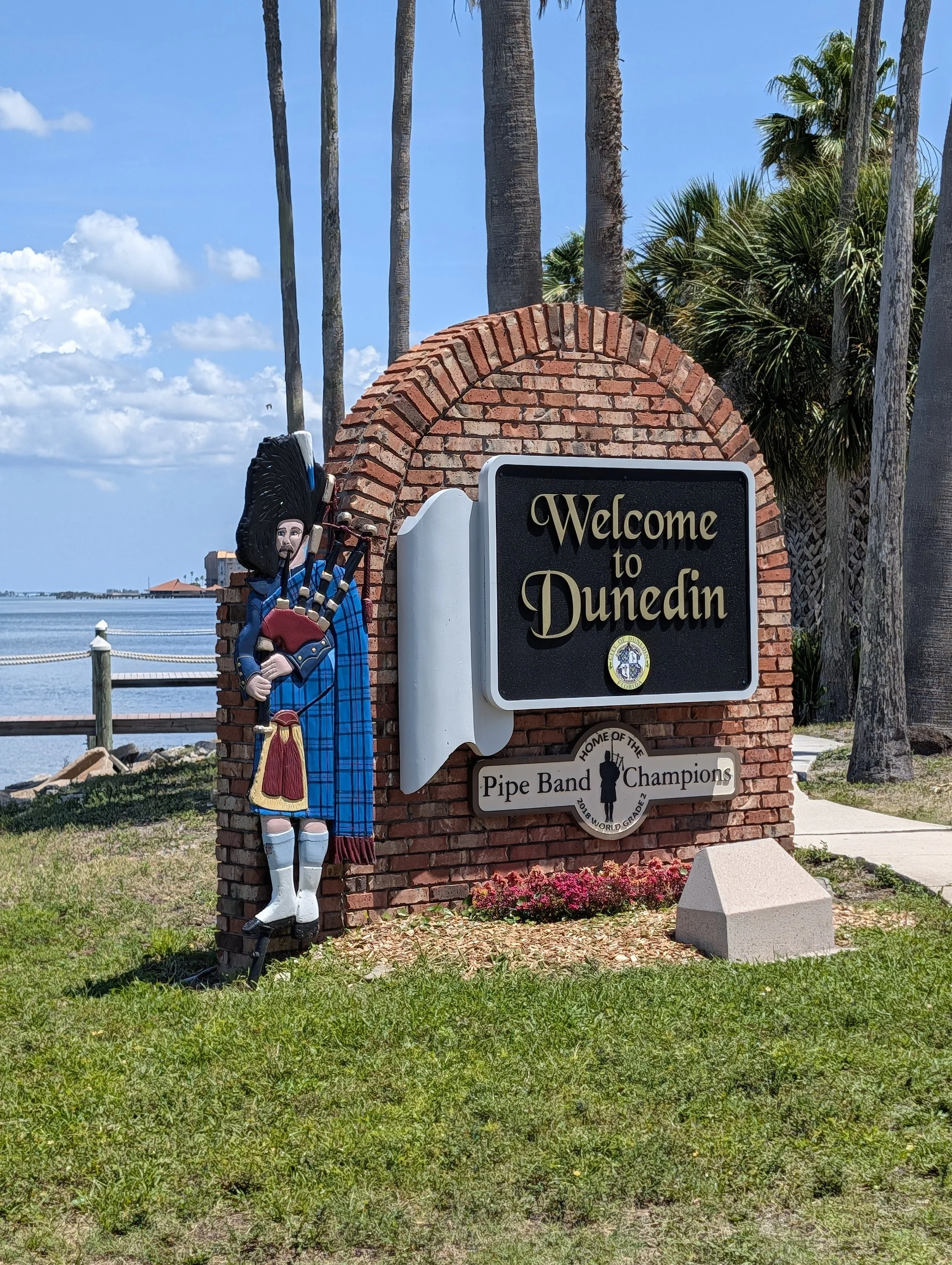 Sign welcoming to Dunedin with a colorful figurine of a bagpiper in traditional Scottish attire, trees, water, and buildings in the background.