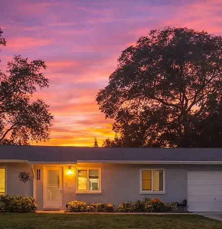Bluebird Cottage in Dunedin, Florida at sunset — a warm and cozy single-story vacation home with illuminated windows, private driveway, and lush greenery, providing the perfect evening retreat after a day touring Dunedin on e-bikes with Paradise Life
