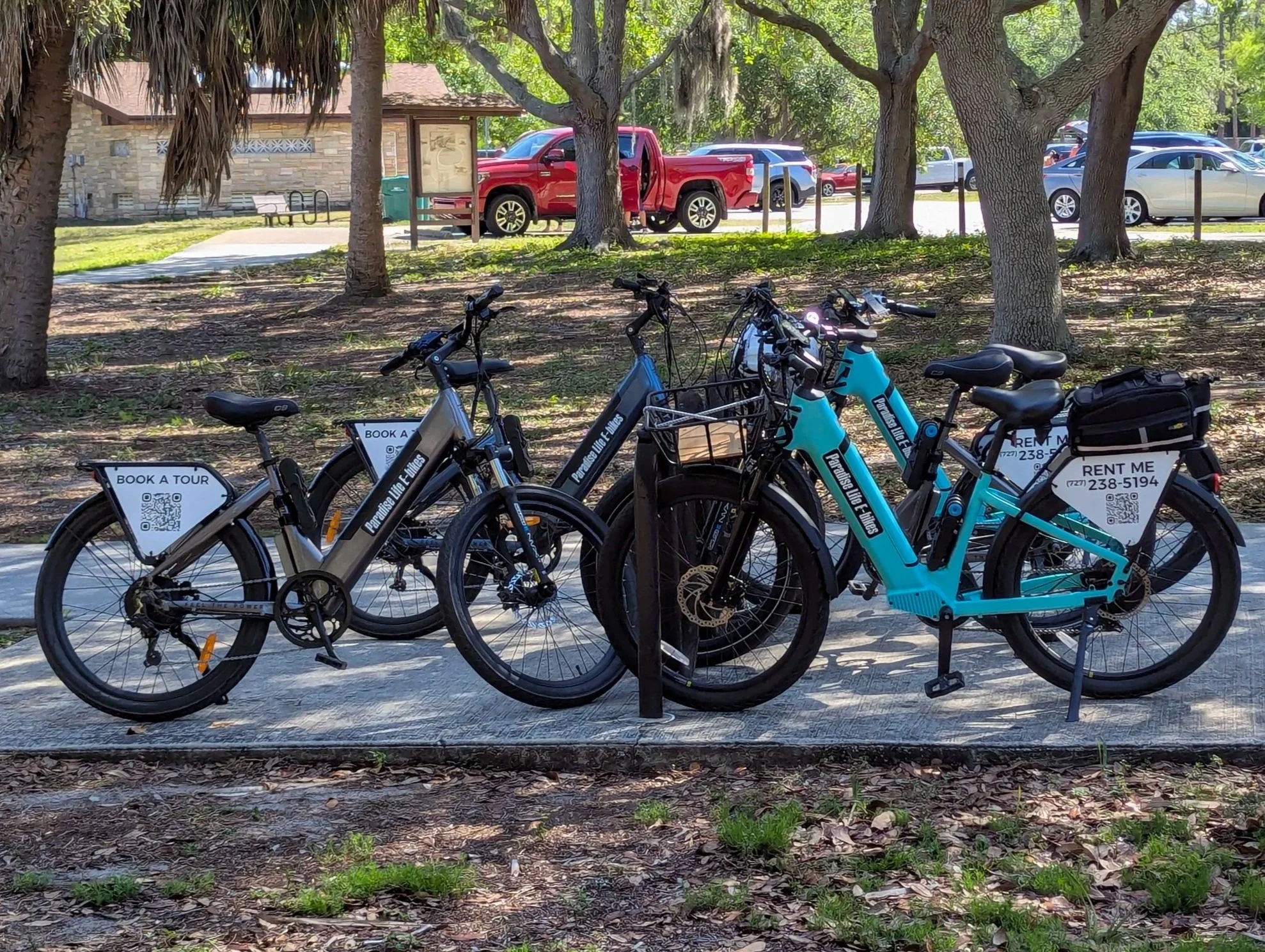 Row of rental bikes parked near a park, with a background of trees, a red truck, and a brick building.