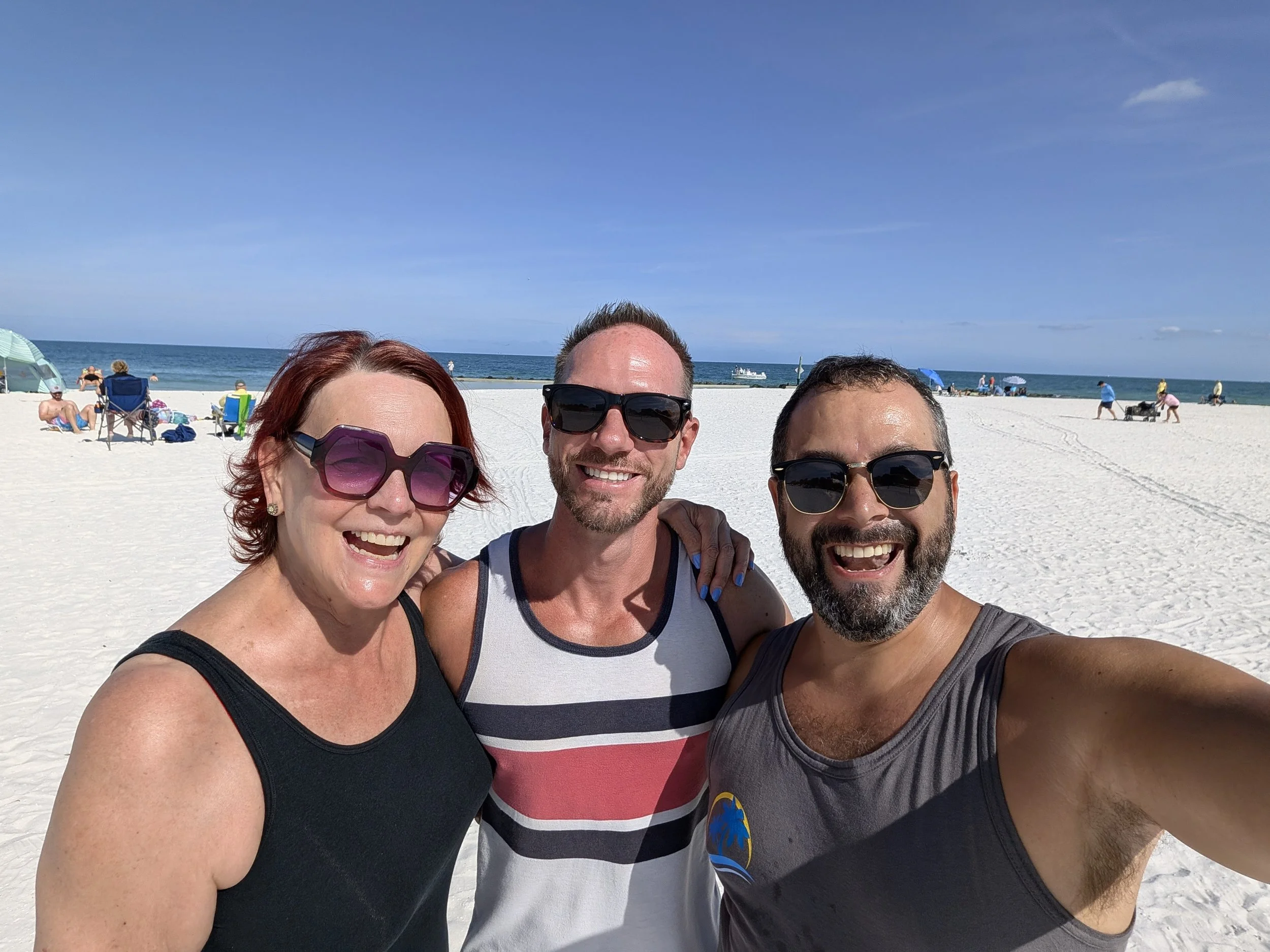 Three smiling people taking a selfie on a beach with white sand, blue sky, and ocean in the background.