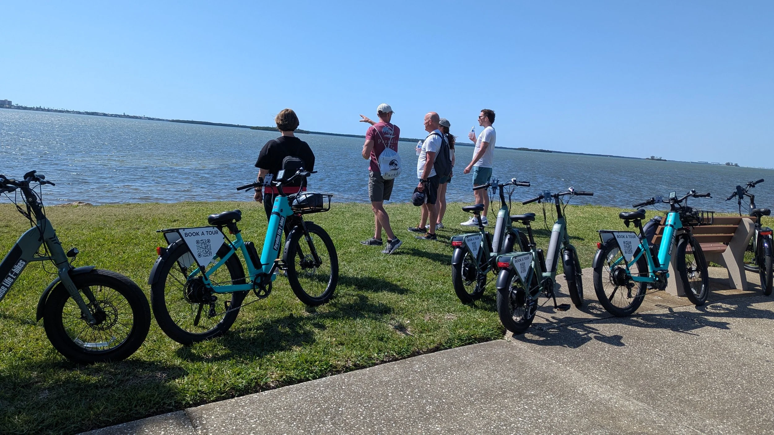 A group of people standing and talking by the water's edge with several bicycles parked nearby, under a clear blue sky.