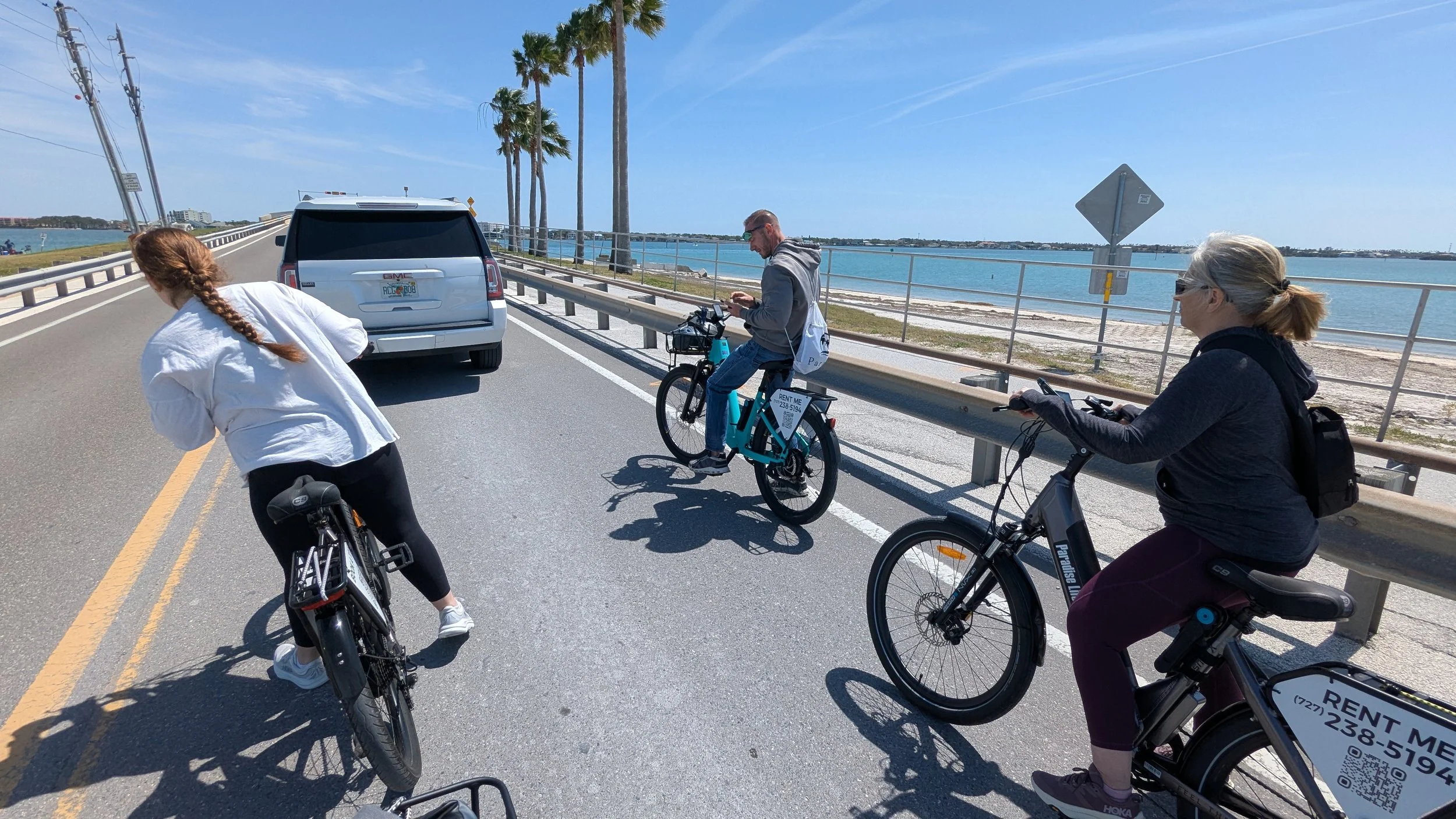 Four people riding bicycles along a coastal road with a water body on the right, palm trees, and a bridge in the background under a blue sky.