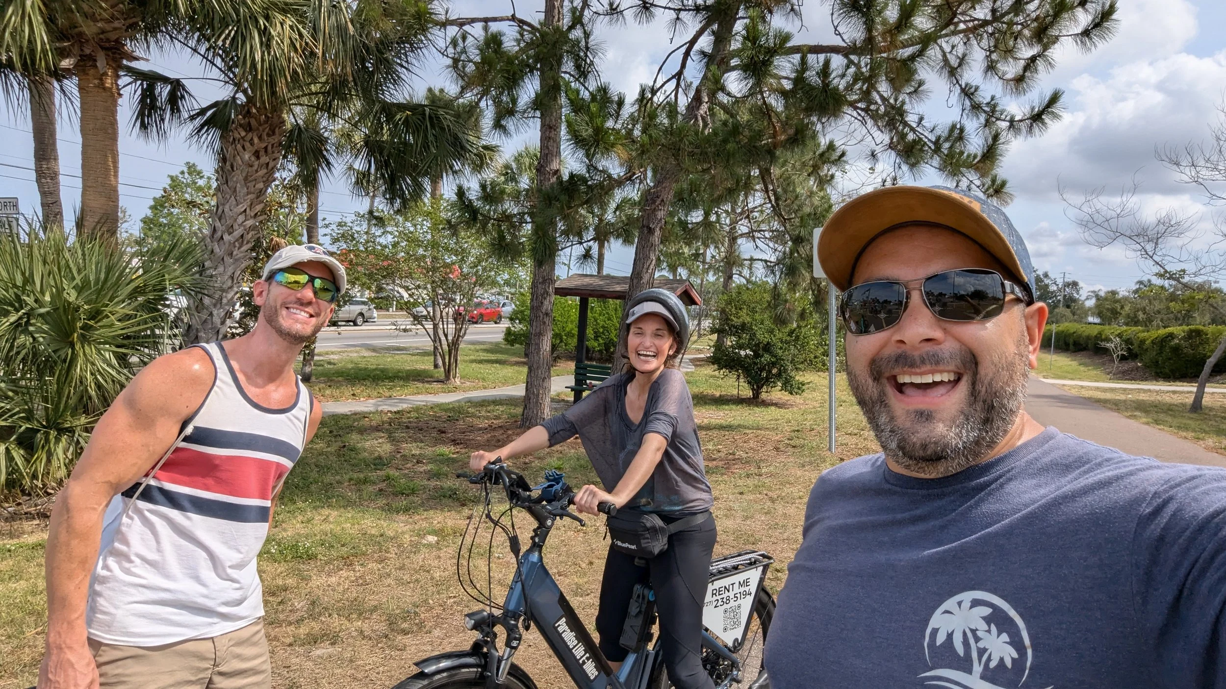 Three people smiling outdoors on a partly cloudy day, one man in a tank top, a woman with a bicycle, and another man in a cap and sunglasses, with trees and a park in the background.