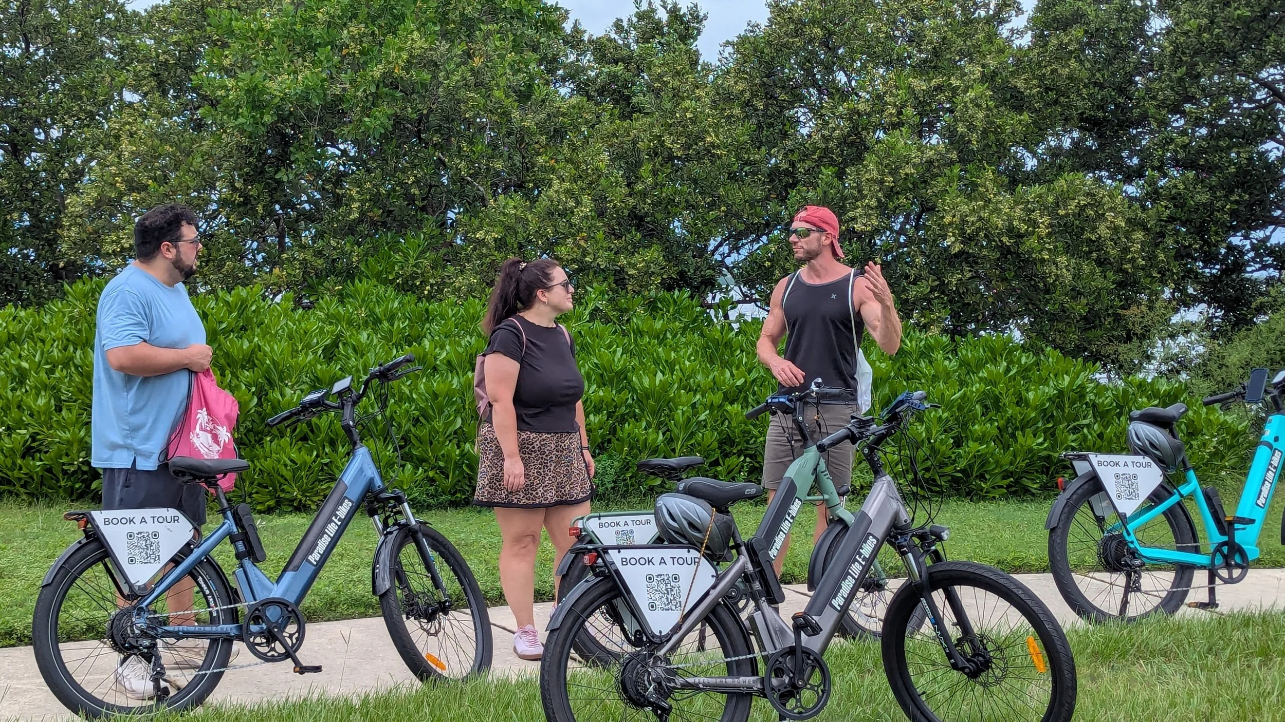 Three people stand outdoors with their bicycles, engaged in conversation. The scene features lush green bushes and trees in the background.