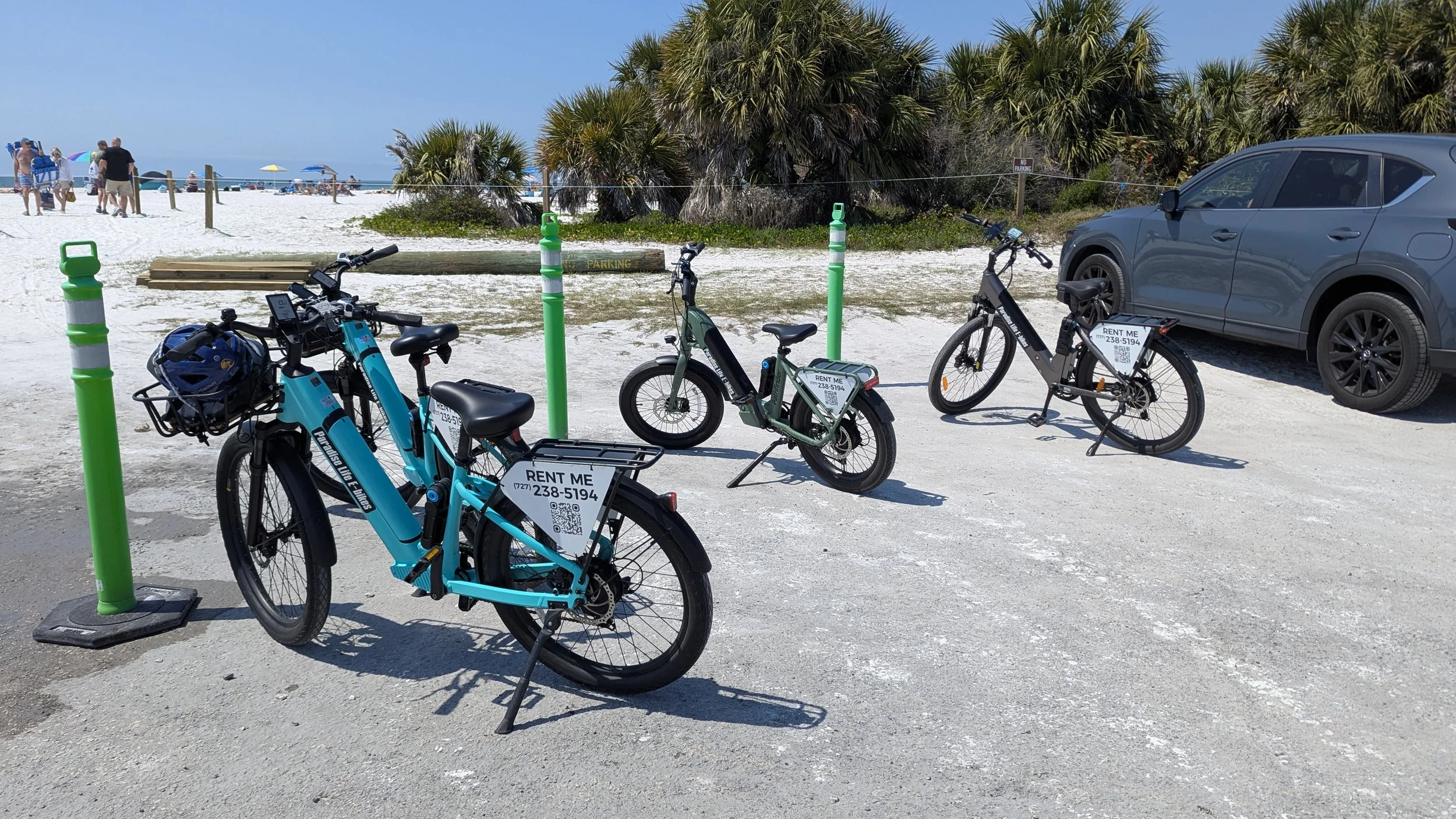 A row of rental bikes parked on a sandy beach parking lot with a gray SUV nearby. The bikes have signs with phone numbers and QR codes for rent. In the background, there are palm trees, a wooden barrier, and people on the beach under umbrellas with t