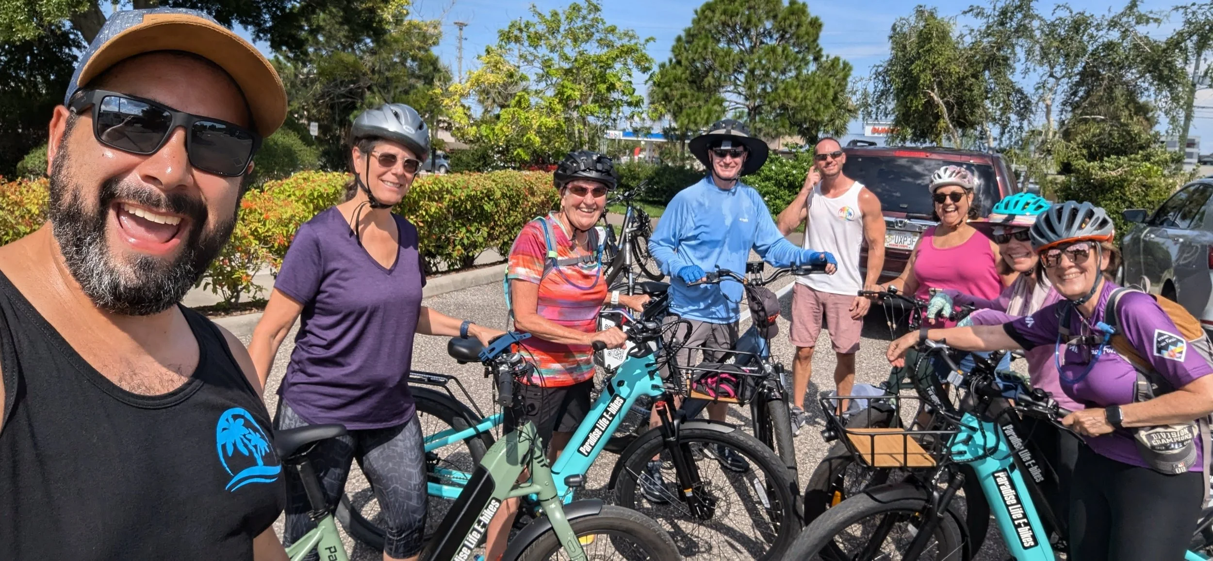 Group of eight people, mostly women, standing with bicycles in a parking lot on a sunny day, wearing helmets and casual sporty clothing, smiling and enjoying a bike ride.