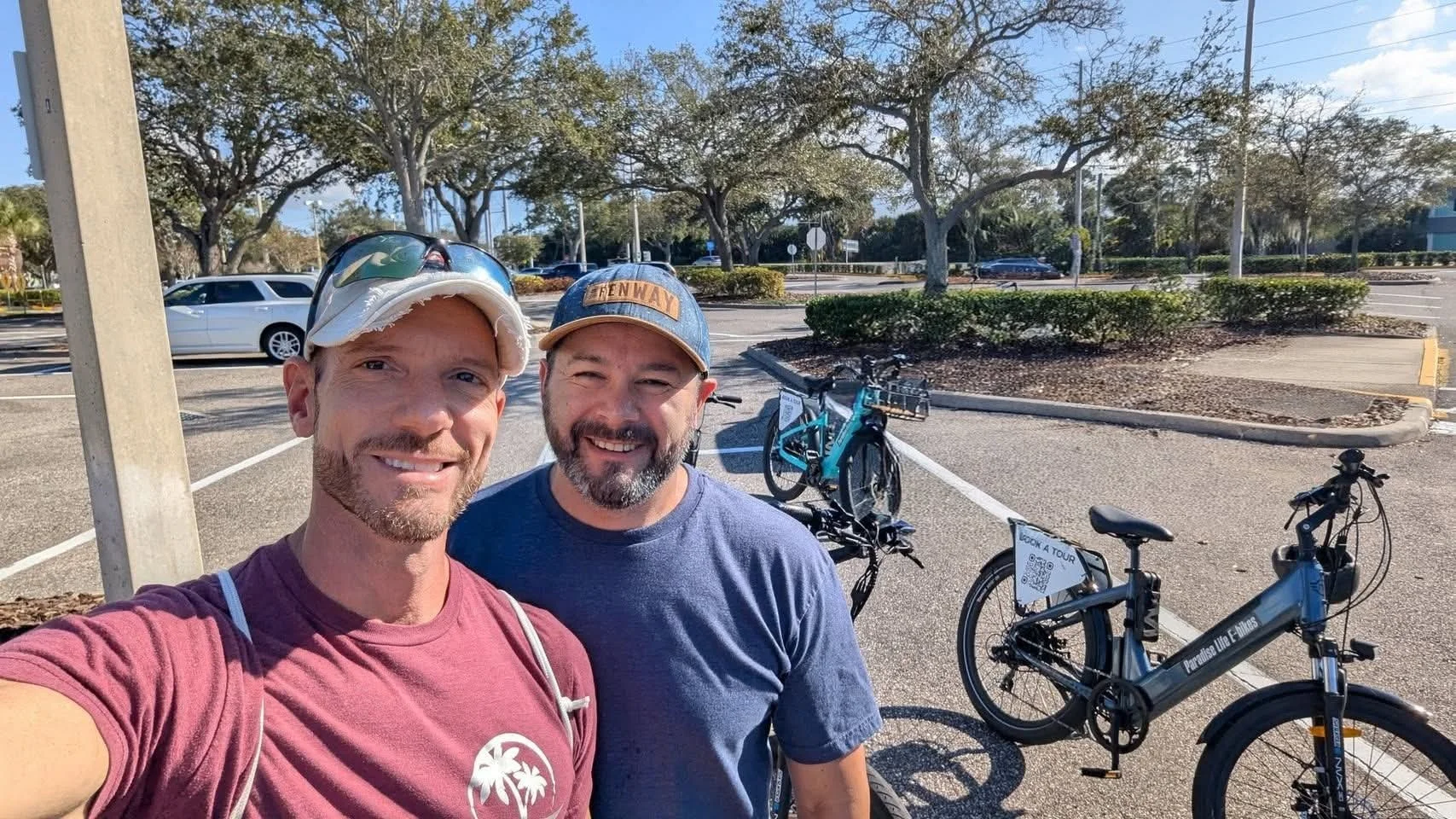 Two smiling men taking a selfie in a parking lot with two bicycles behind them, trees, and a clear blue sky in the background.