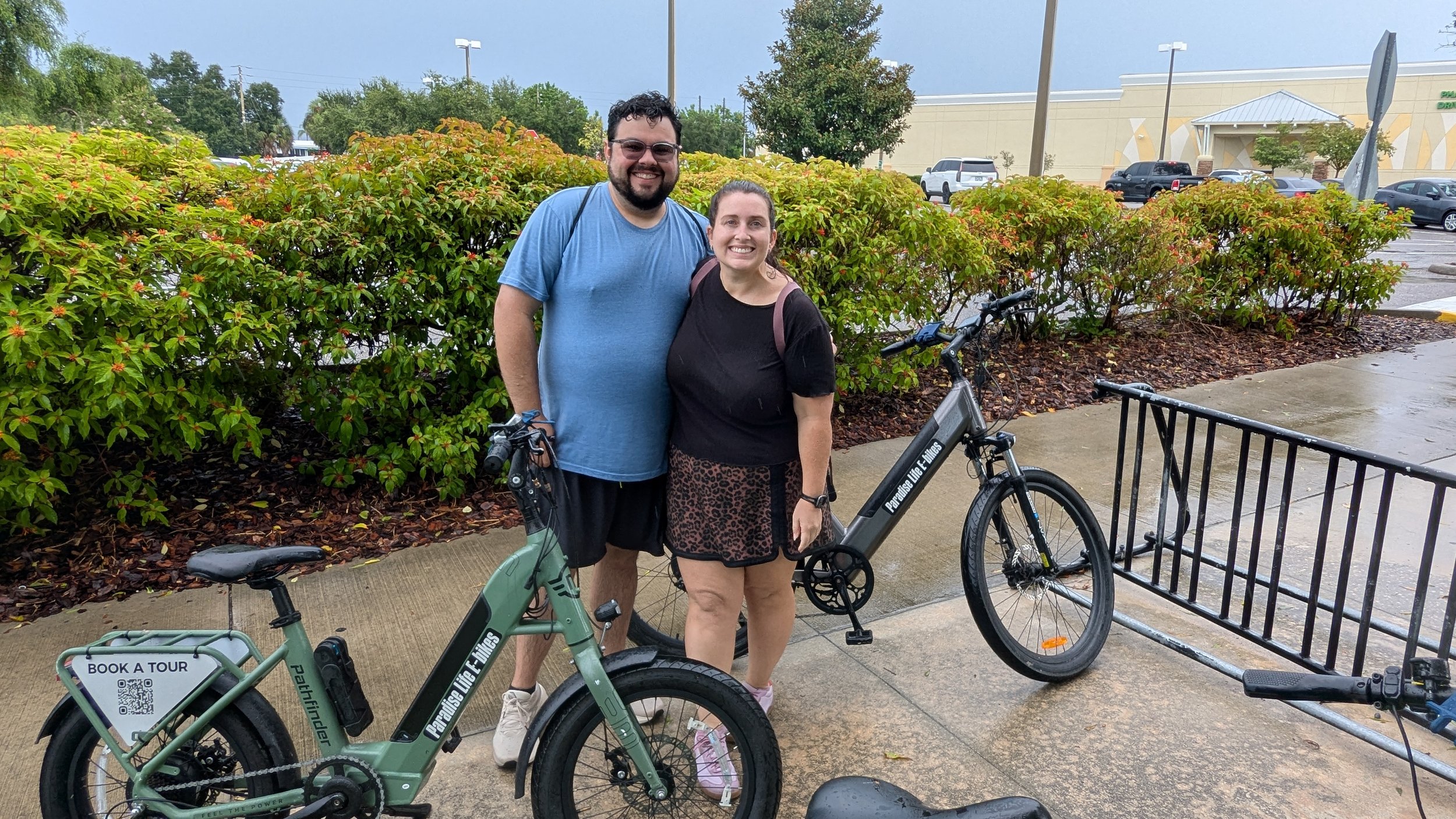 A man and a woman smiling outdoors, standing next to electric bikes, in a parking lot near bushes and a shopping mall on a rainy day.