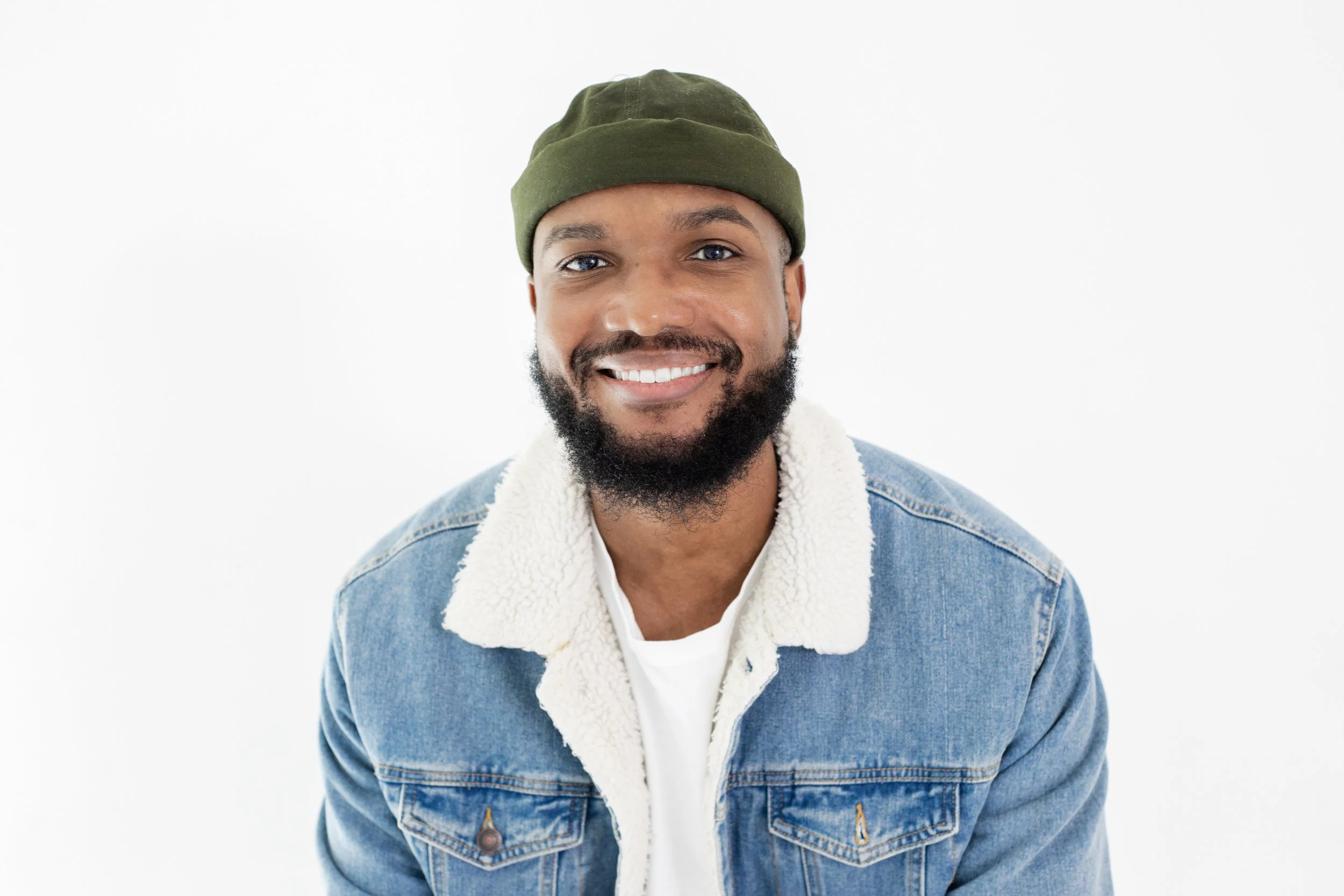 Smiling young man with a beard wearing a green beanie, denim jacket with a shearling collar, and a white shirt, against a plain white background.