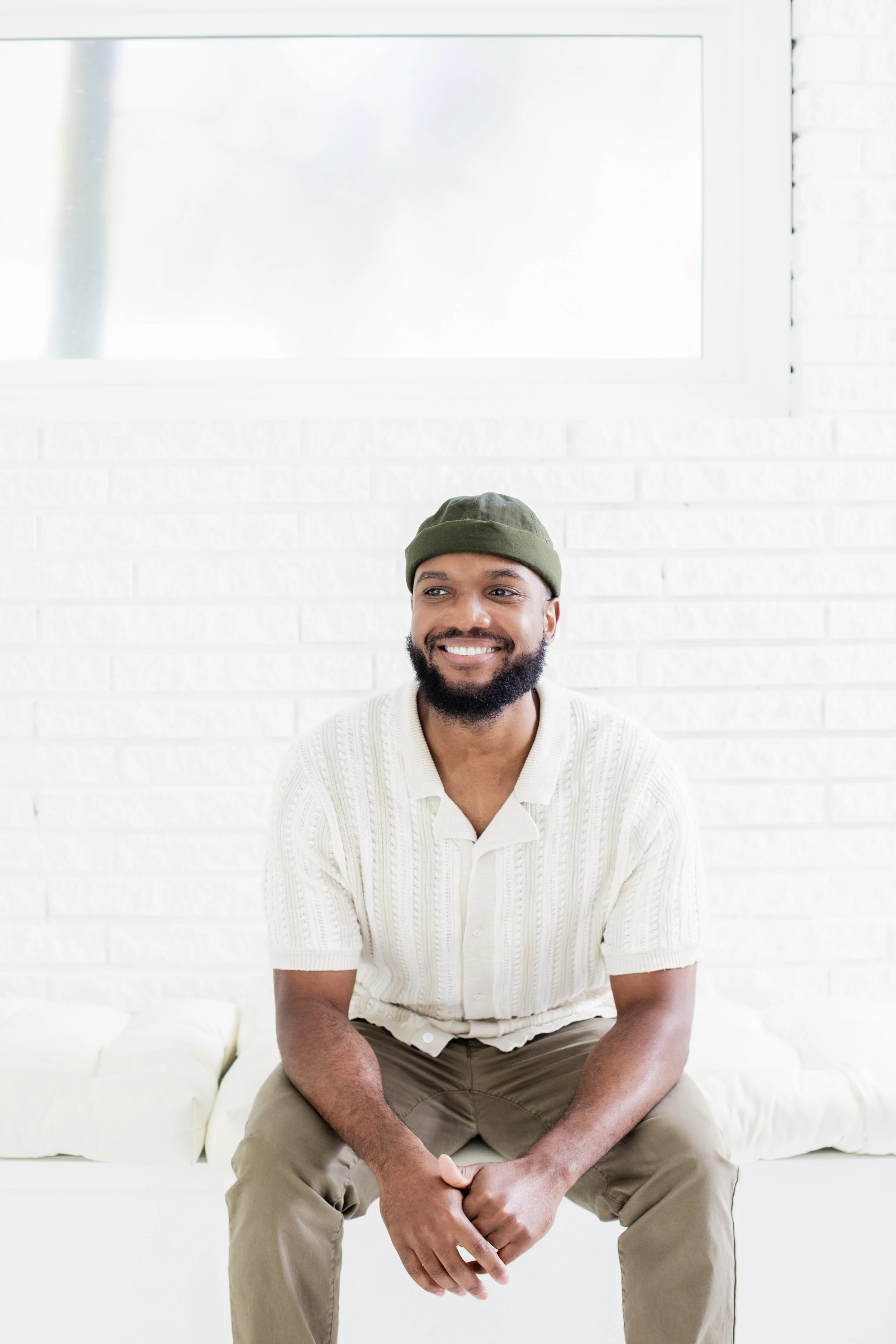 A smiling man sits on a white sofa in a room with white brick walls and a large window.