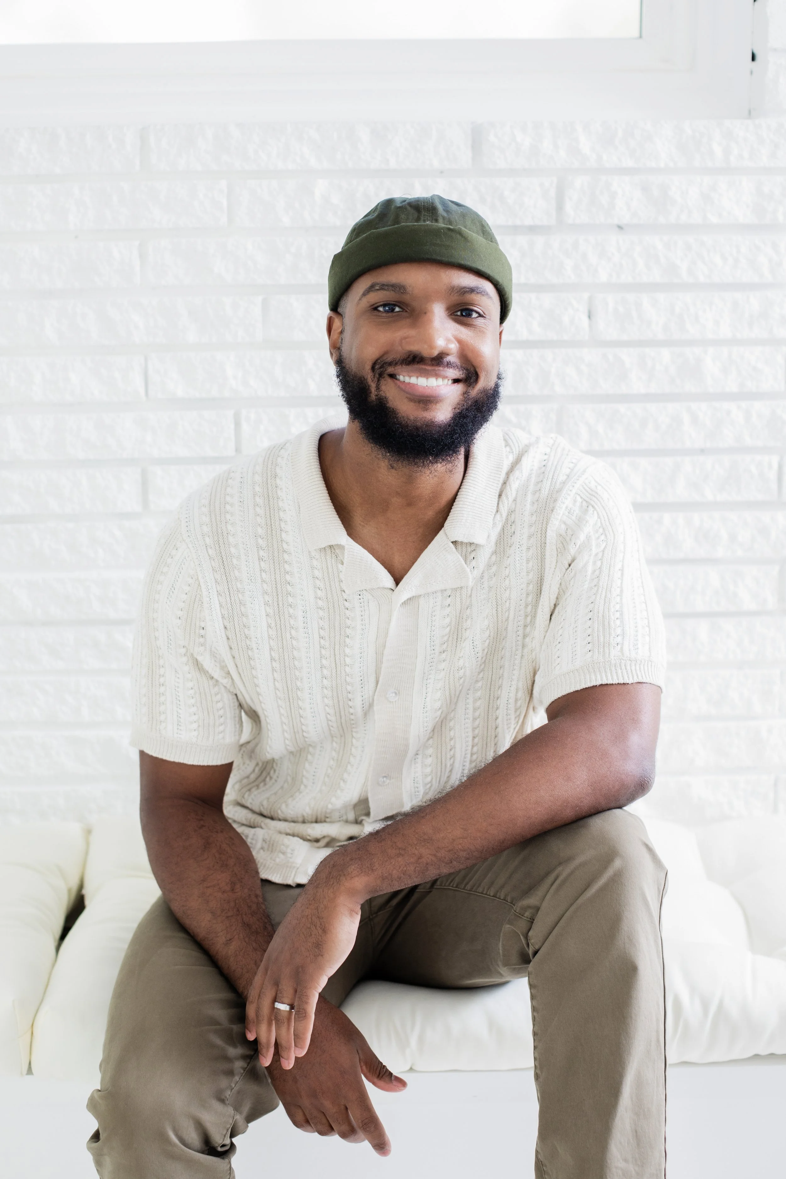 A smiling man sitting on a white couch with a white brick wall background, wearing a white short-sleeved polo shirt, khaki pants, and a green beanie.