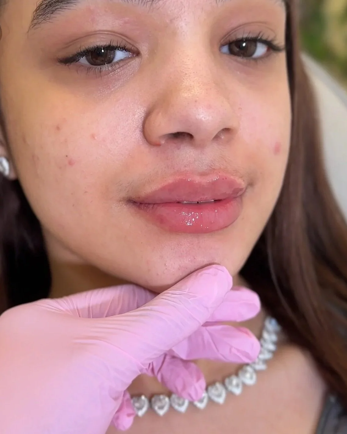 Close-up of a young woman with brown eyes and long brown hair, wearing a pearl necklace. A gloved hand is gently touching her chin.