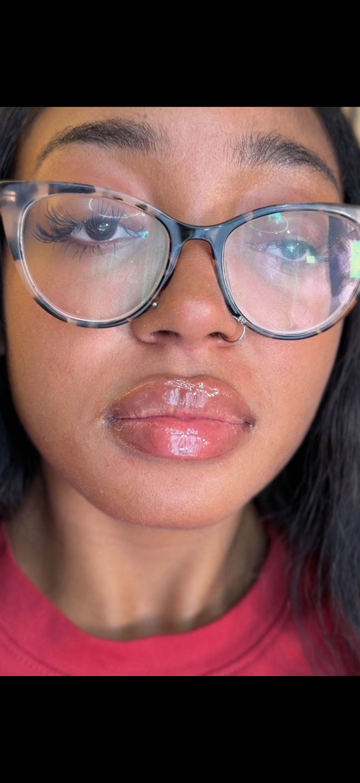 Close-up of a woman wearing oversized glasses with reflections in the lenses, glossy lips, and a nose ring, dressed in a red top.