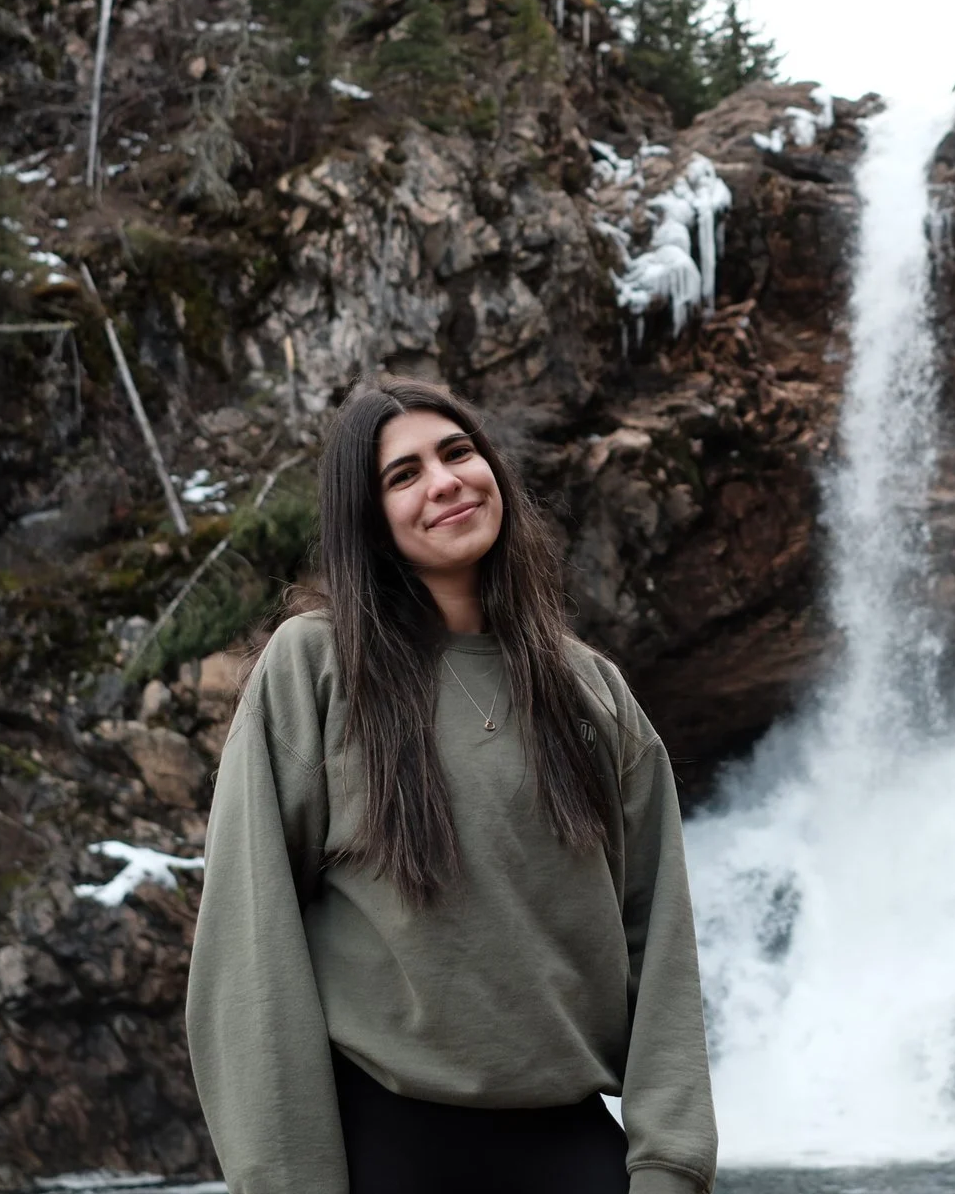 A young woman with long dark hair standing in front of a waterfall with ice formations on rocks and some snow.