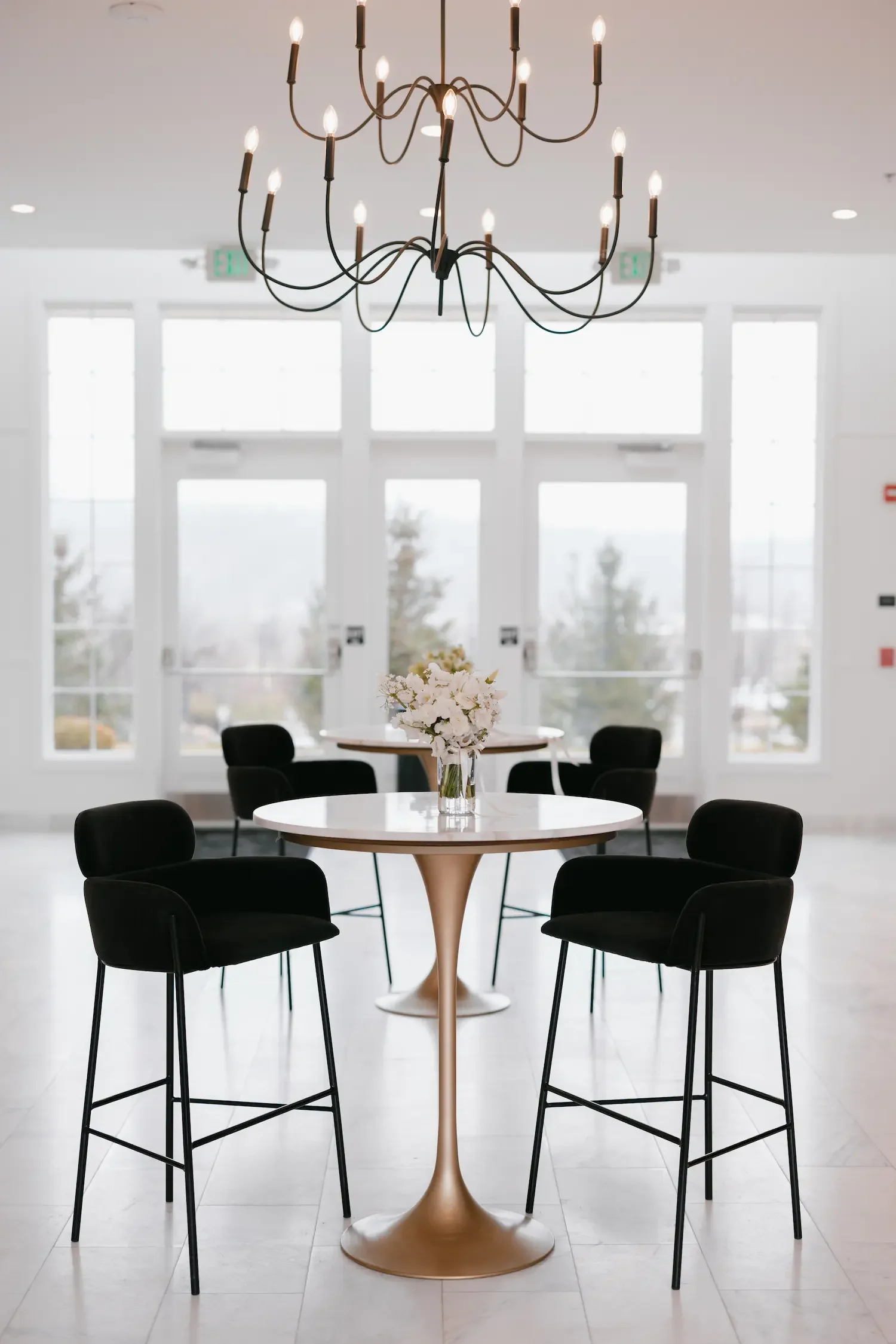 Modern dining room with a round table decorated with a vase of white flowers, surrounded by six black chairs, large windows in the background, and a black chandelier hanging from the ceiling.
