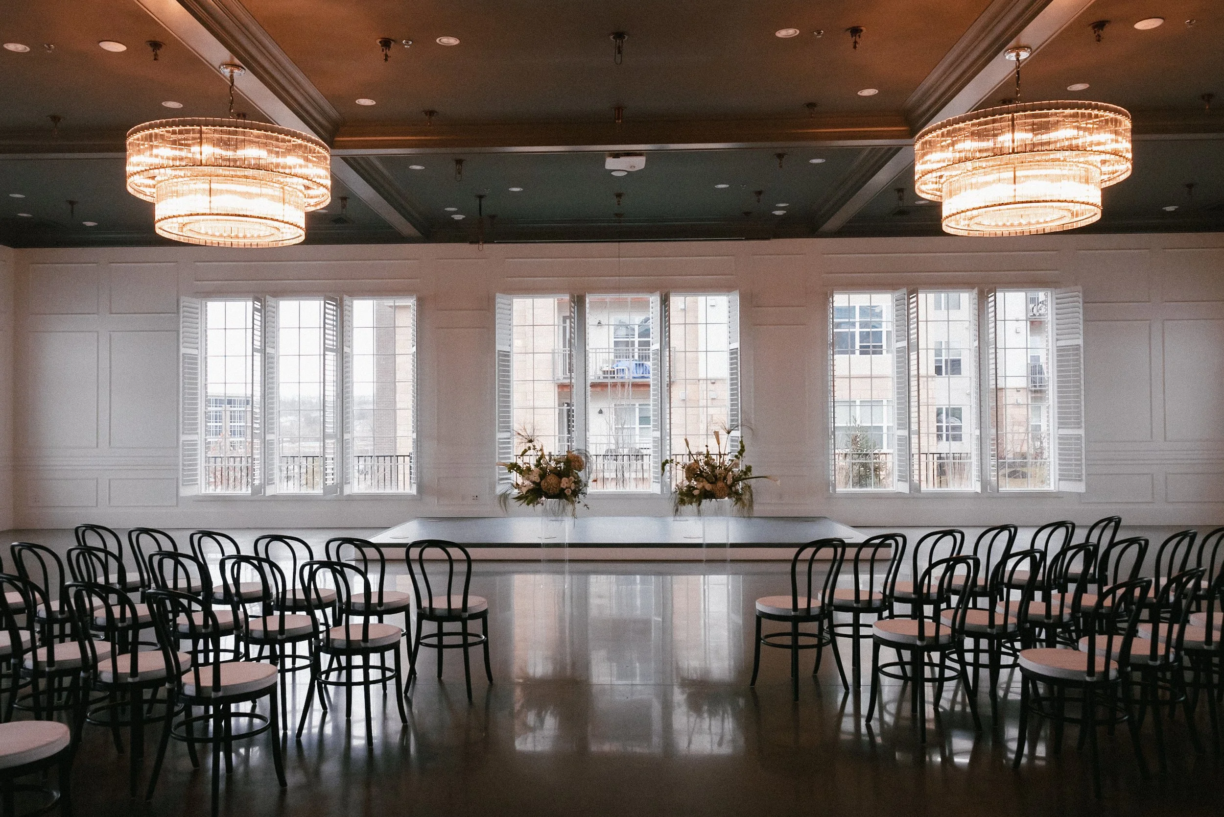 Empty event room with black chairs around a rectangular table, large windows with shutters, and chandeliers hanging from the ceiling.