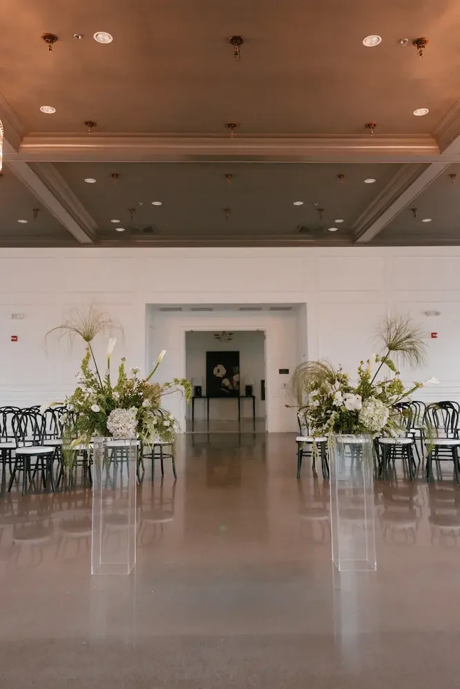 An elegant event hall with white walls, a polished floor, and floral arrangements on tall, transparent stands. The hall has rows of black chairs and a doorway leading to another room.